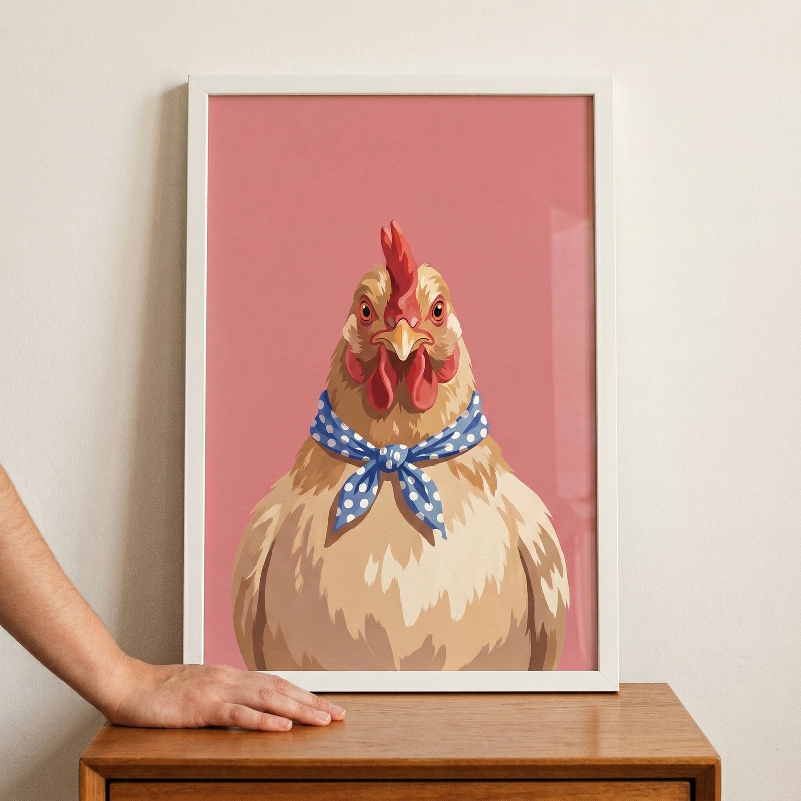 Blue polka dot hen wall art in a white frame on a wooden table with a hand touching it against a pink background.
