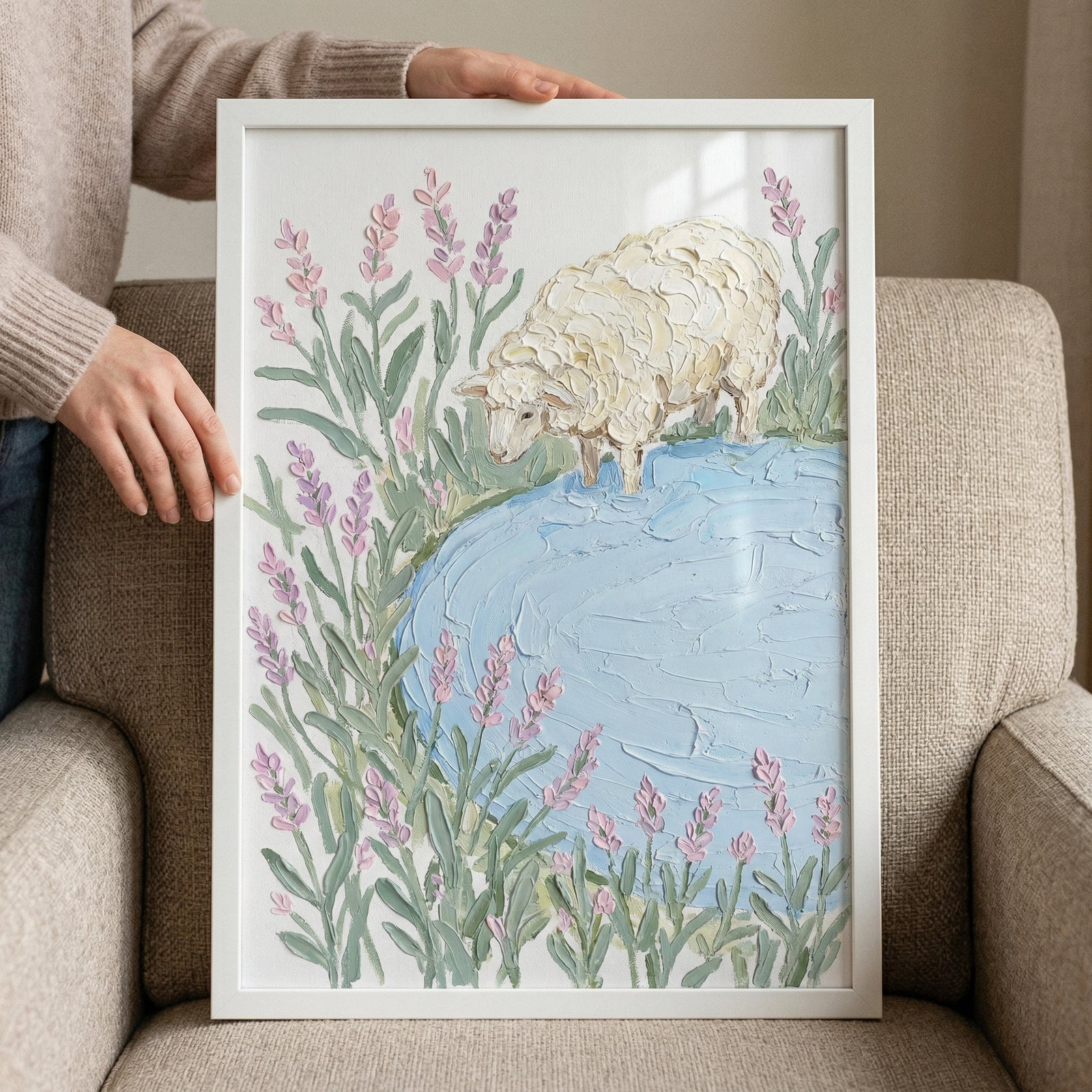 Framed painting of a textured white sheep standing at the edge of a soft blue pond surrounded by tall lavender stems rests against a cushioned chair while hands hold the frame. The pastel colors and thick brushstrokes create a calm gentle countryside scene.
