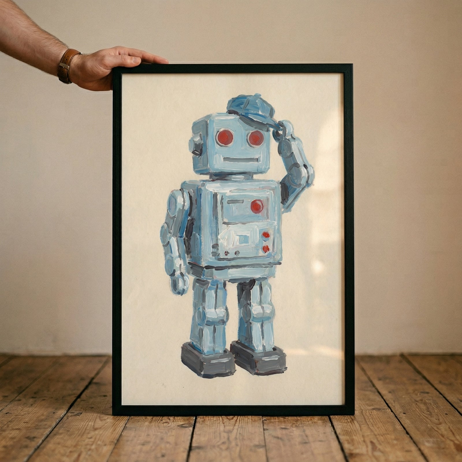 A person's hand, wearing a brown watch, holds the top edge of a black rectangular frame. Within the frame is a piece of art depicting a vintage-style blue robot. The robot has a square head with two round red eyes and is tipping a small blue cap with its right hand. 