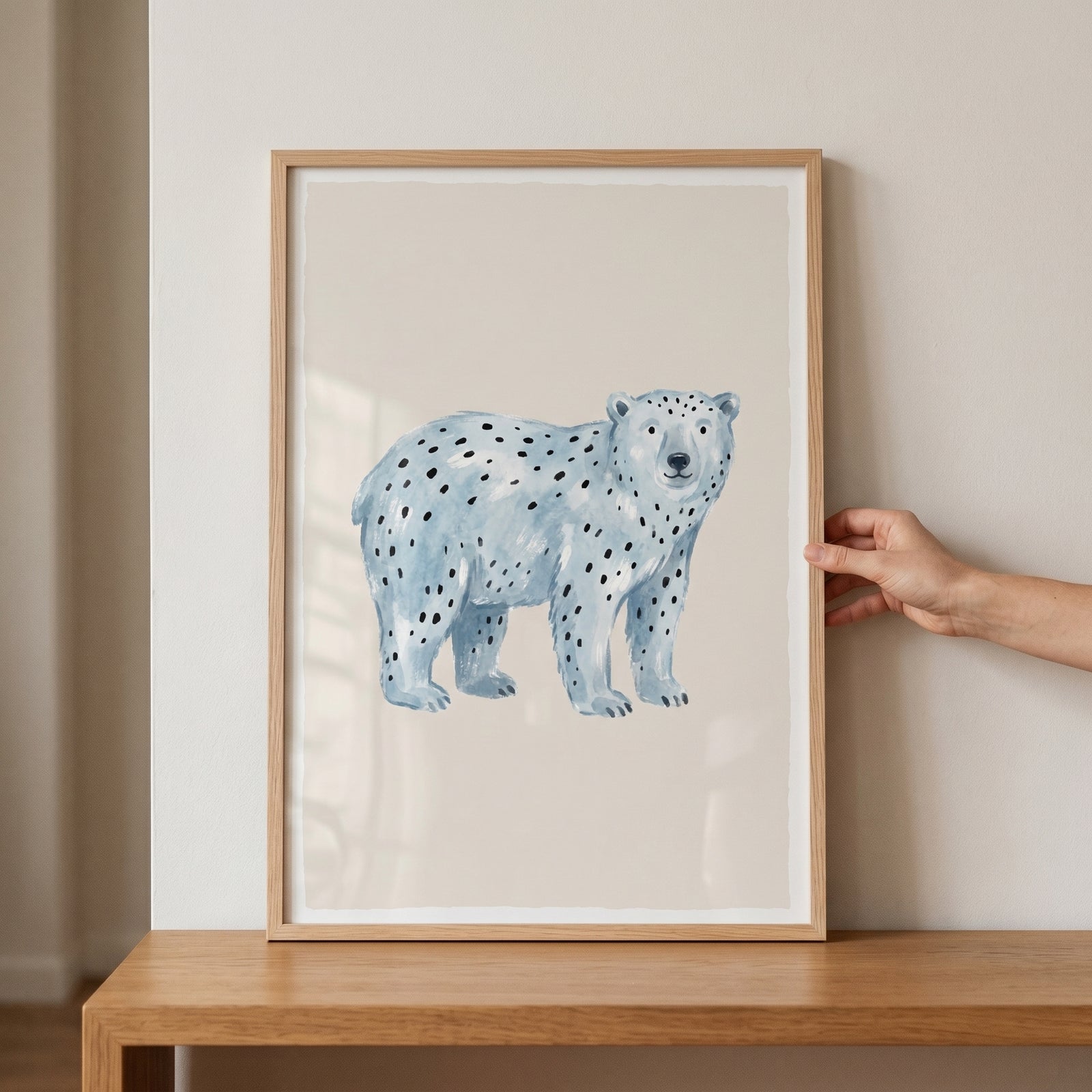 Framed blue spotted polar bear wall art resting on a wood shelf against a neutral wall, held by a hand.