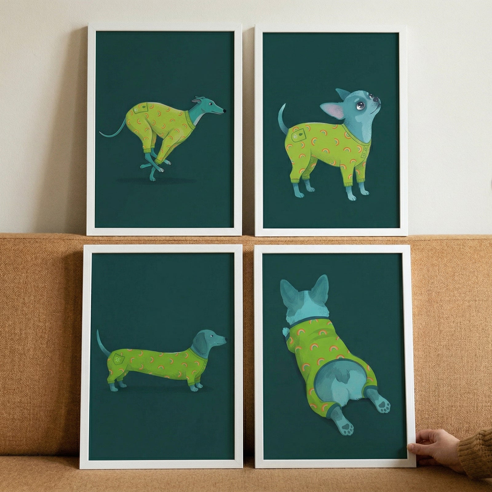 Four framed illustrations are arranged in a square grid on a light brown fabric surface. Each illustration features a stylized blue dog wearing bright green pajamas decorated with a pattern of colorful rainbows and small pockets. The dogs are set against a solid dark teal background.
