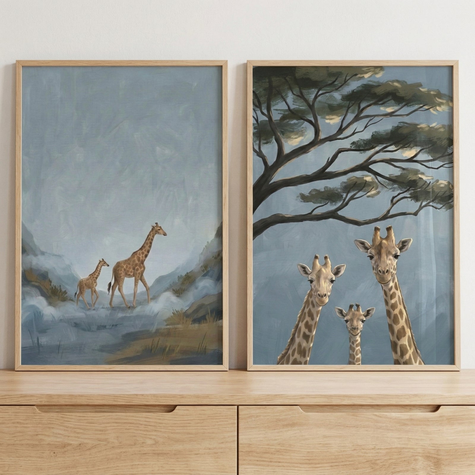 Two framed giraffe paintings rest on a light wood dresser against a white wall. One shows an adult giraffe walking with a calf through a misty blue landscape, while the other shows three giraffes looking forward beneath spreading tree branches against a soft blue sky.