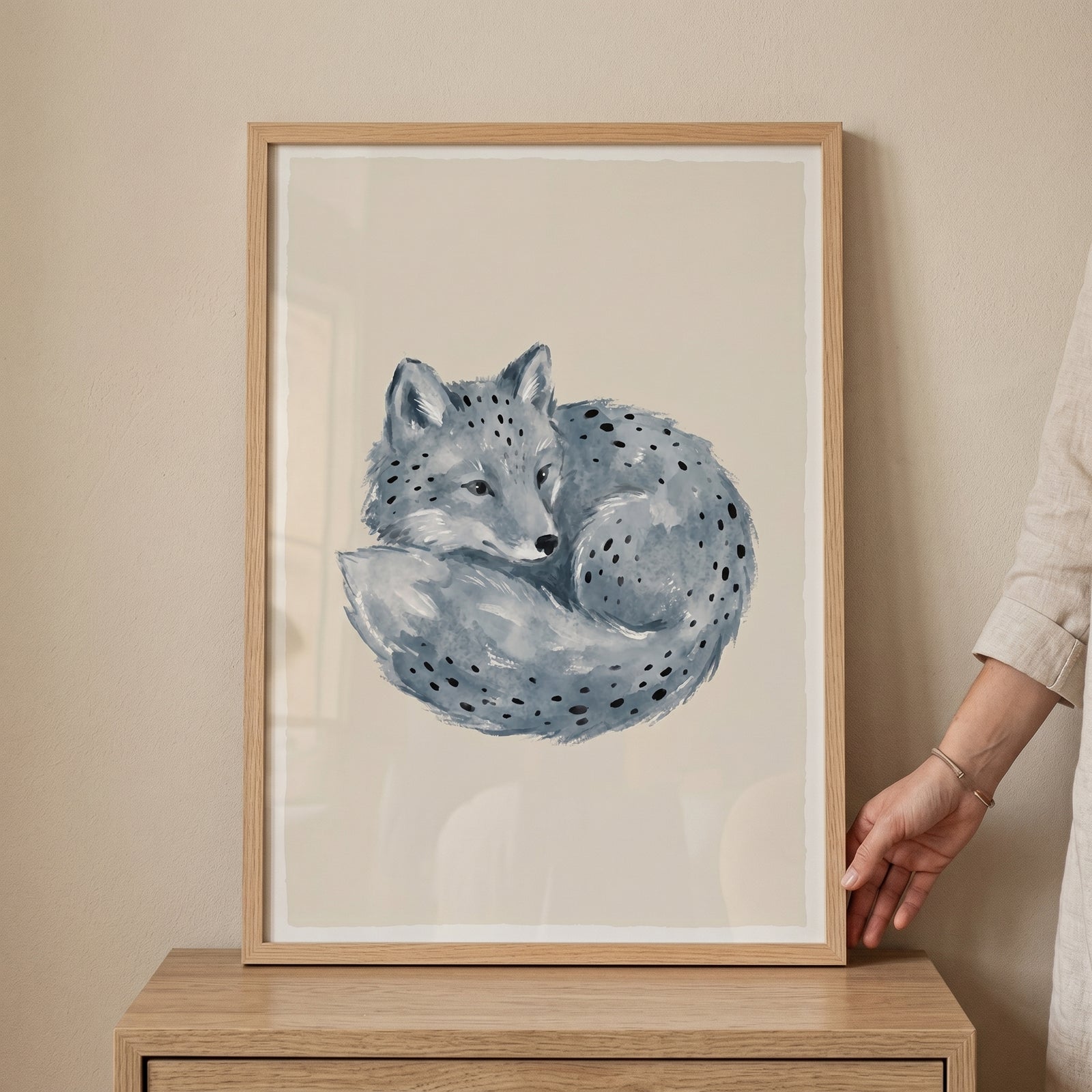 Framed blue watercolor curled fox wall art rests on a wooden dresser while a hand touches the frame.