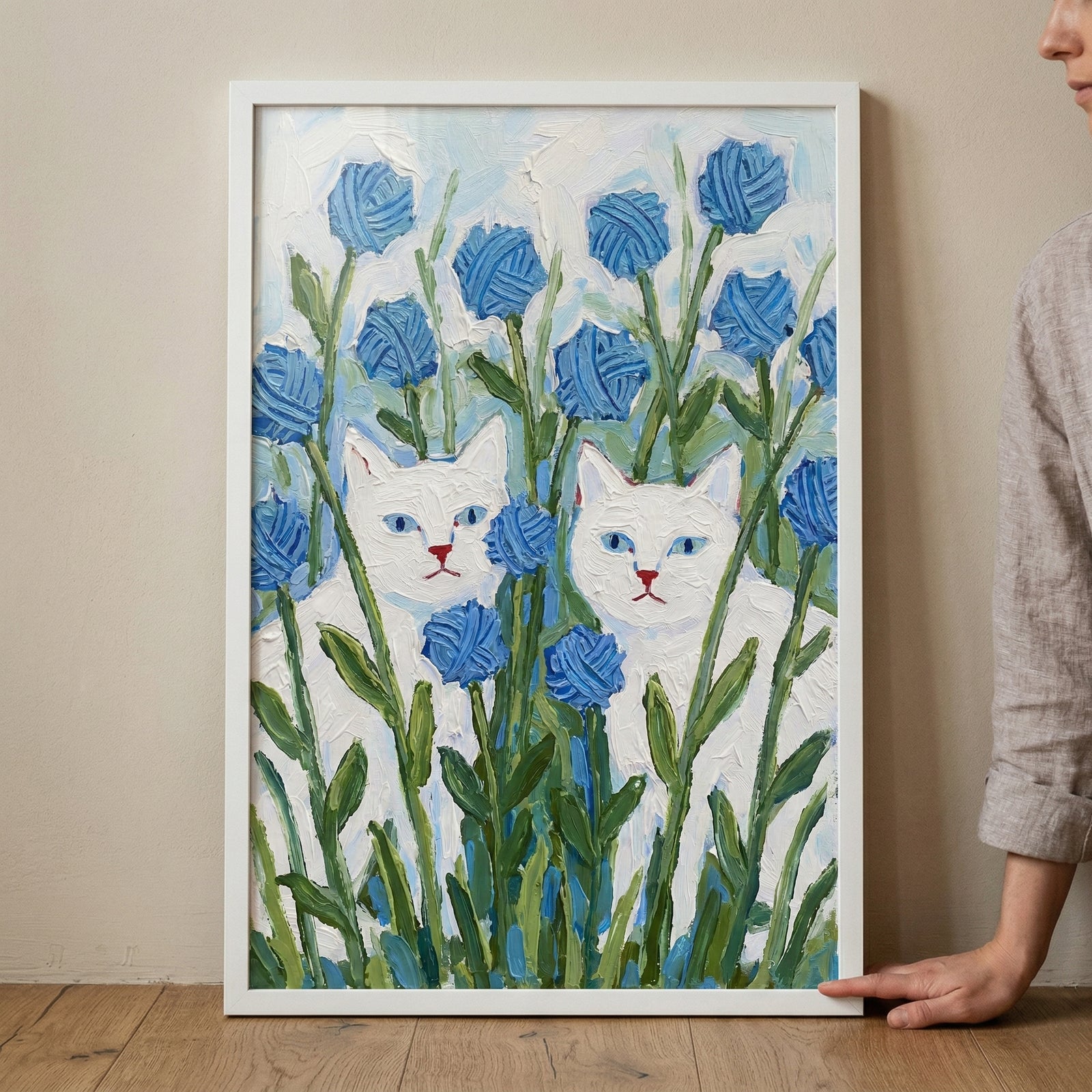 A framed vertical artwork depicts two white cats with vivid blue eyes nestled amongst tall green stalks and round, textured blue flowers. The entire piece is created with thick, expressive applications of color, giving the scene a tactile feel. The cats have simple, charming features with small red-pink noses. Behind them, the sky is a wash of light blue and white. The artwork is held in a slim white frame, placed on a light wooden floor against a neutral wall.