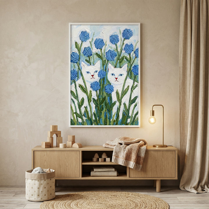 Framed painting of two white cats with blue eyes peeking through tall green stems and round blue flowers, displayed above a wooden cabinet in a softly lit neutral room. A small lamp, folded knit blanket, and wooden toys surround the artwork, making it a calm focal point in the space.