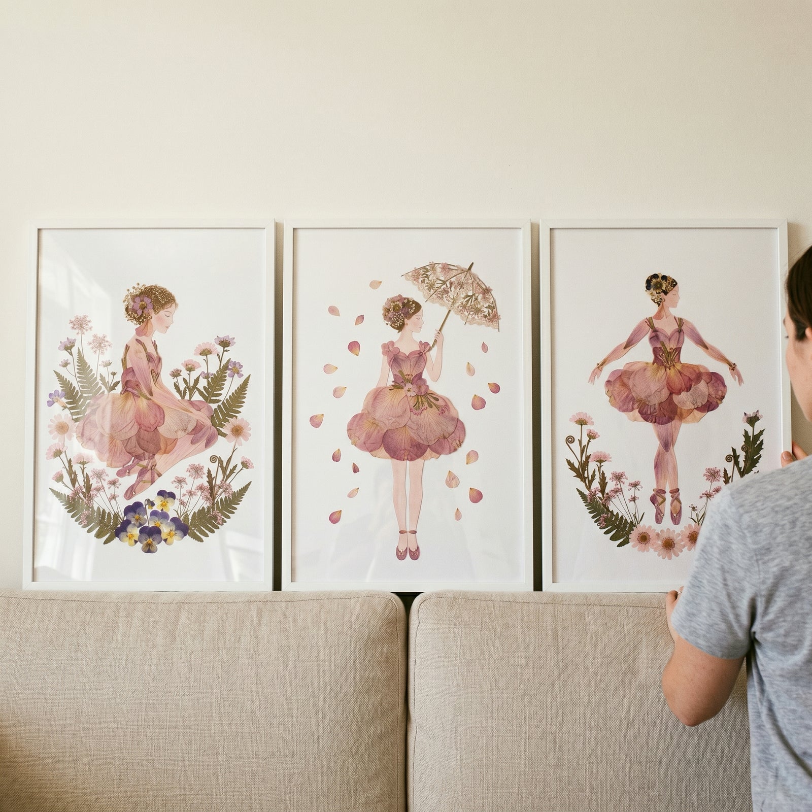 Blush botanical flower girl wall art set of 3 in white frames above a beige sofa as a person adjusts the third frame.