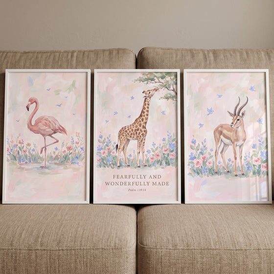 Blush botanical safari animals wall art set of 3 in white frames with a flamingo, giraffe, and gazelle on a tan couch.