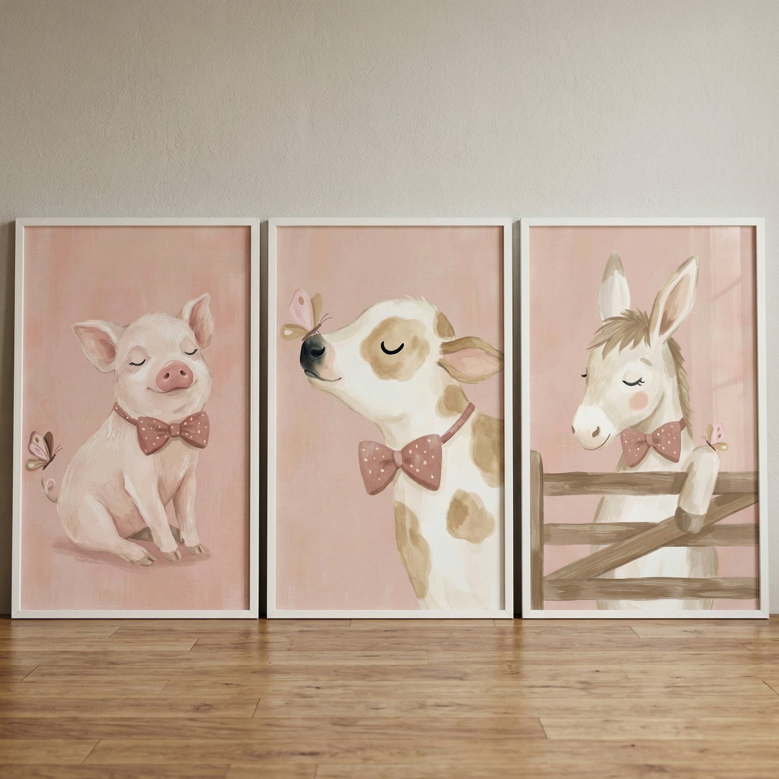 Three white-framed rectangular illustrations are displayed side-by-side against a neutral wall, resting on a light wood-grained floor. Each illustration features a soft, pale pink background and a gentle, whimsical animal subject.

The left-hand illustration shows a small, pale pink pig with its eyes closed in a content expression, wearing a darker pink bow tie with white polka dots. A tiny butterfly floats near the pig's curly tail.