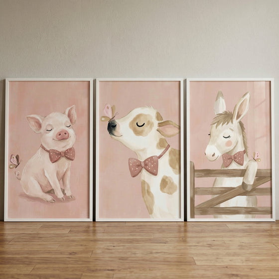 Three white-framed rectangular illustrations are displayed side-by-side against a neutral wall, resting on a light wood-grained floor. Each illustration features a soft, pale pink background and a gentle, whimsical animal subject.

The left-hand illustration shows a small, pale pink pig with its eyes closed in a content expression, wearing a darker pink bow tie with white polka dots. A tiny butterfly floats near the pig's curly tail.