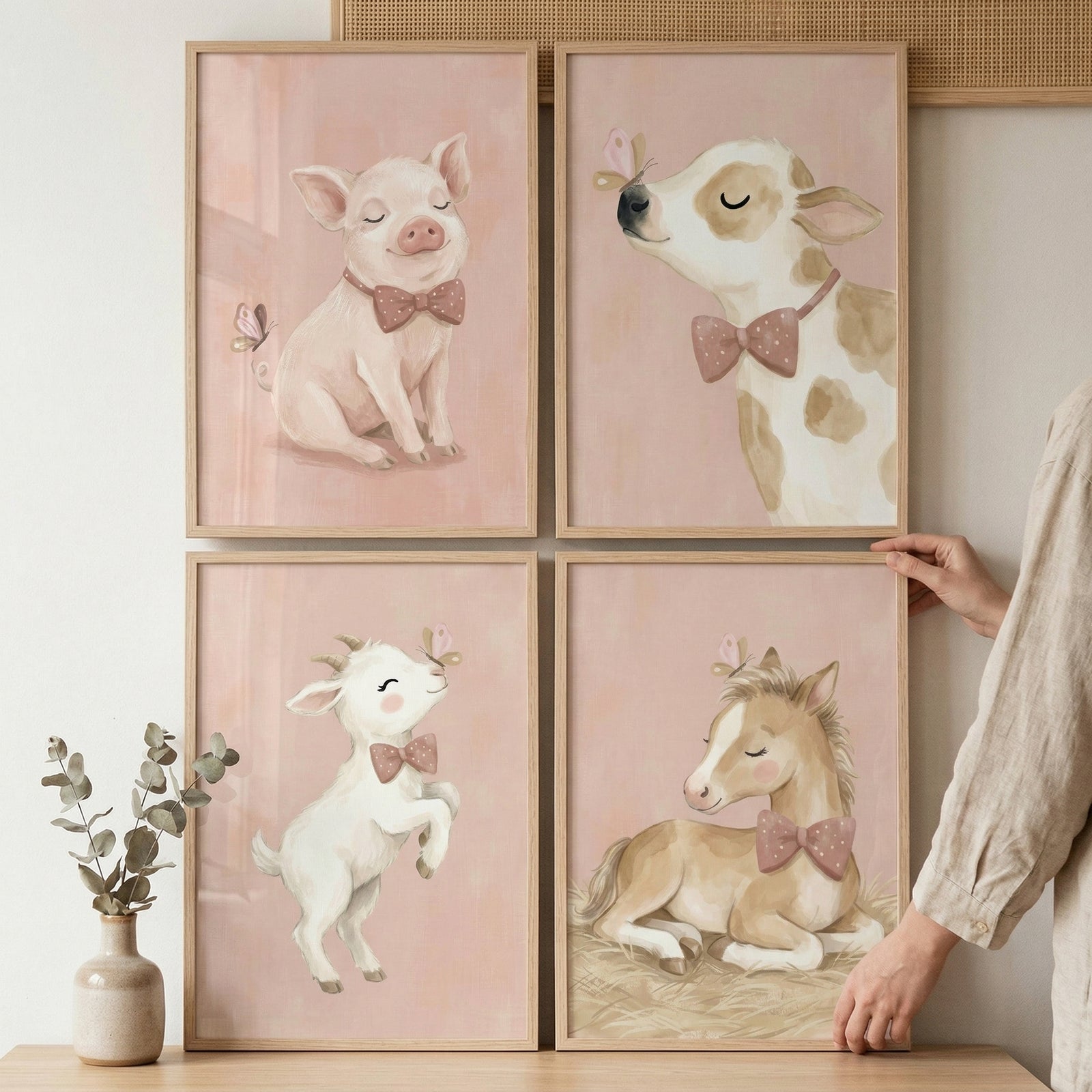 This set of four framed illustrations features soft, whimsical depictions of young farm animals, each set against a muted pink background and encased in a light-colored wooden frame. The four pieces are arranged in a square grid on a wall.
