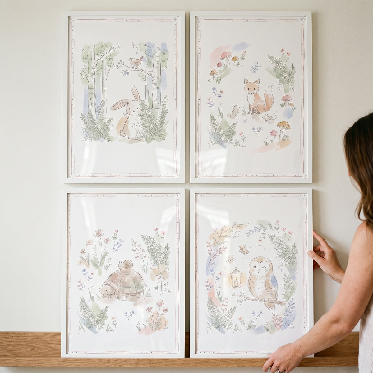A person adjusts a blush floral forest animal wall art set of 4 featuring a bunny, fox, turtle, and owl in white frames.