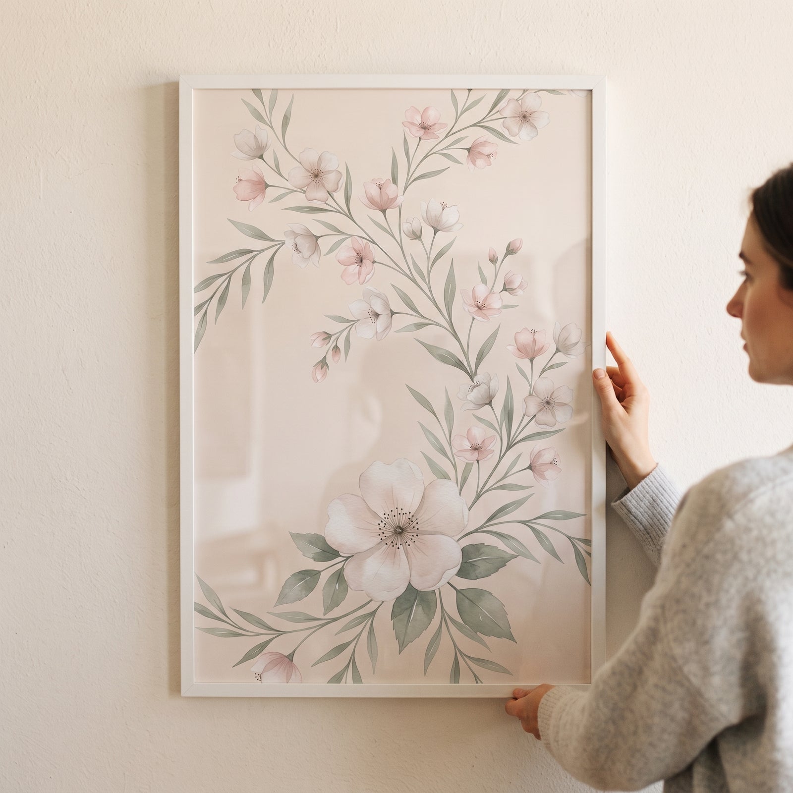 A woman holds a framed blush pink botanical floral print with delicate watercolor blossoms and soft green leaves.