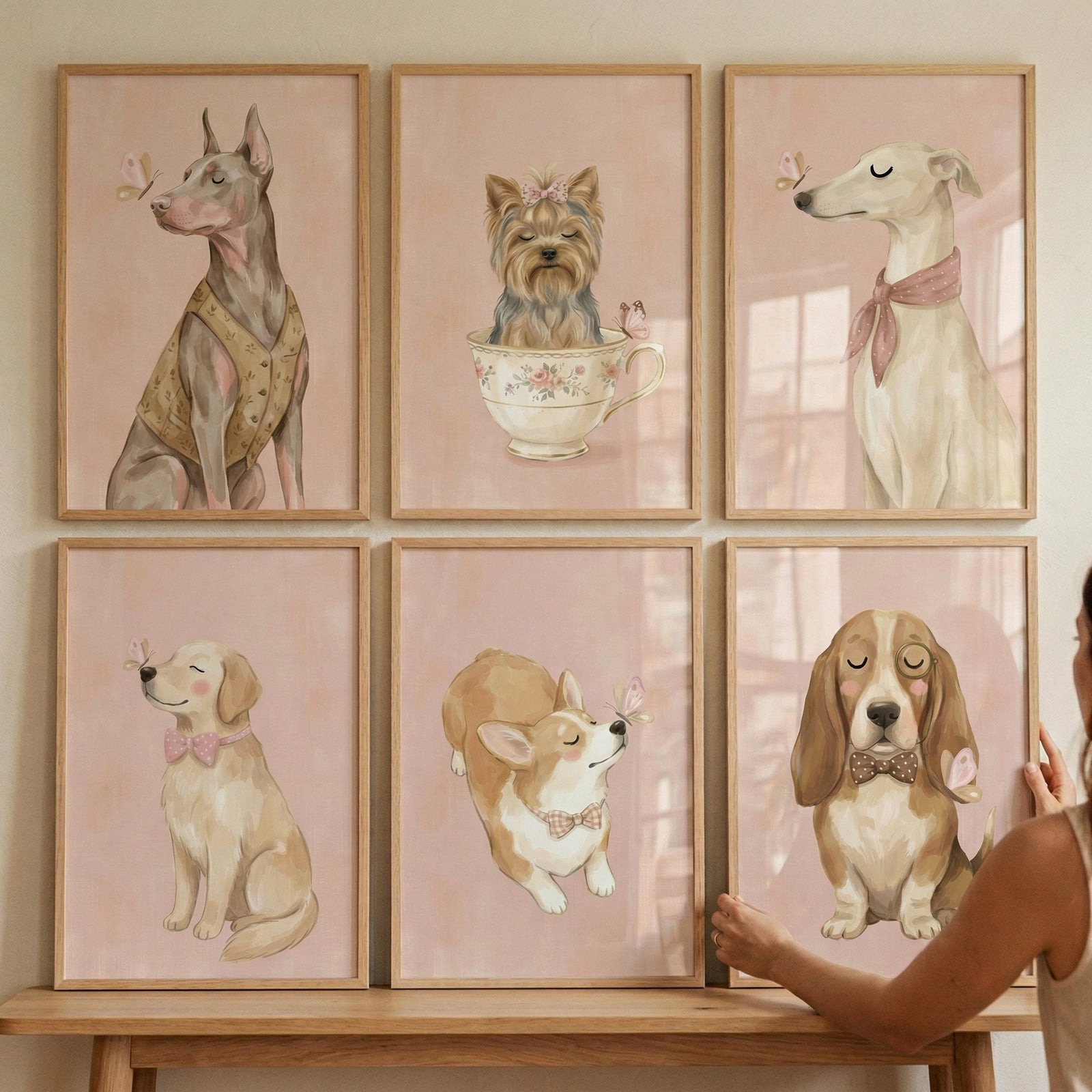 A set of six framed illustrations of various dog breeds displayed in a grid on a wall. The artwork is in a soft, whimsical digital style with a gentle pastel color palette of pink, tan, and cream. Each frame features a different dog: a slender greyhound wearing a pink scarf, a small terrier sitting inside a floral teacup, a sleek dog in a patterned vest, a golden puppy with a pink bow tie, a corgi with a decorative collar, and a long-eared hound wearing a bow tie.