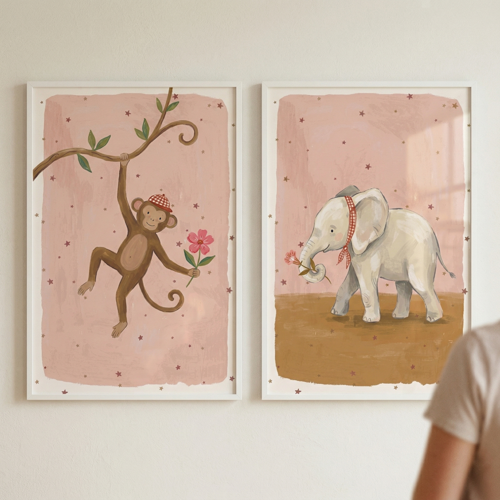 Two framed illustrations on a soft pink star patterned background hang side by side on a wall. One shows a smiling monkey wearing a red checkered hat swinging from a tree branch while holding a pink flower, and the other shows a small elephant with a matching headband gently holding a flower in its trunk while a bird perches nearby.
