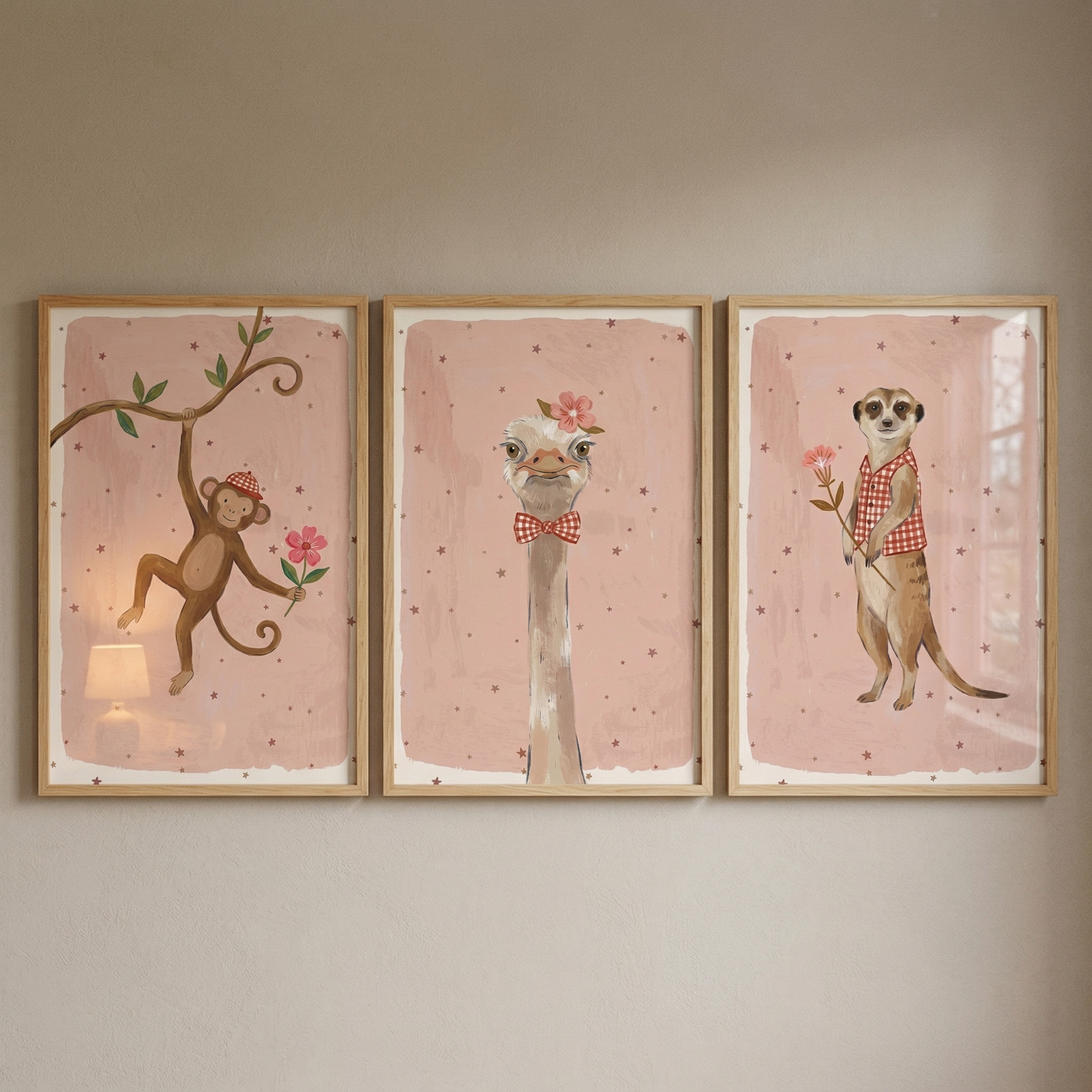 Three rectangular framed vertical artworks hang side-by-side on a plain, neutral wall. Each illustration features a whimsical animal against a soft pink background dotted with small, dark stars. The far-left piece shows a brown monkey hanging from a leafy branch, wearing a red-and-white checkered cap and holding a pink flower. 