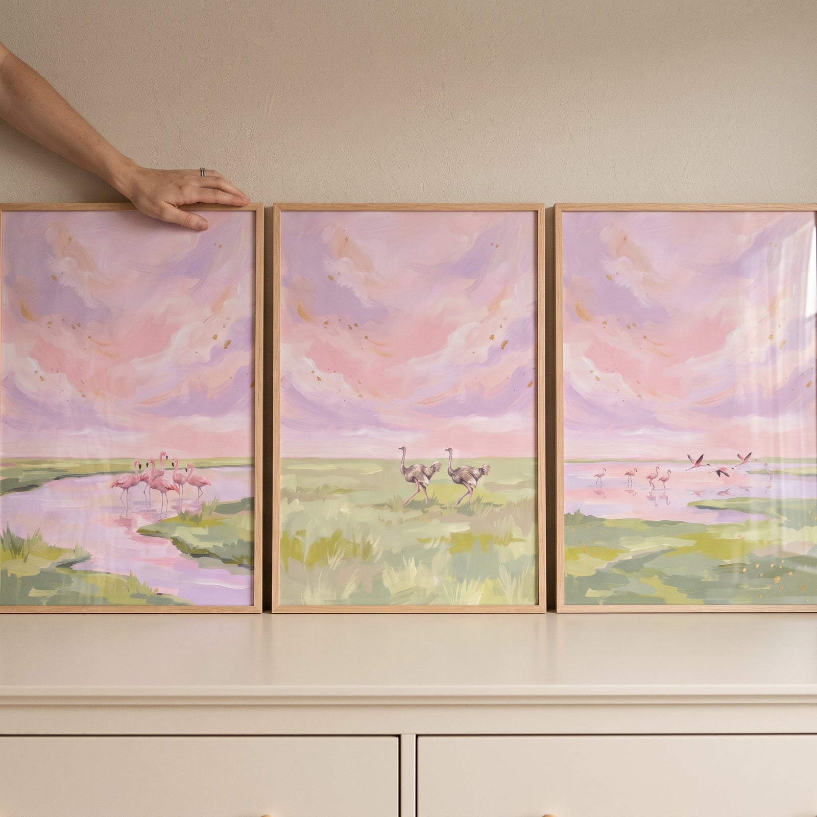 A hand touches a blush pink savanna birds wall art set of 3 showing flamingos and ostriches in a pastel landscape.
