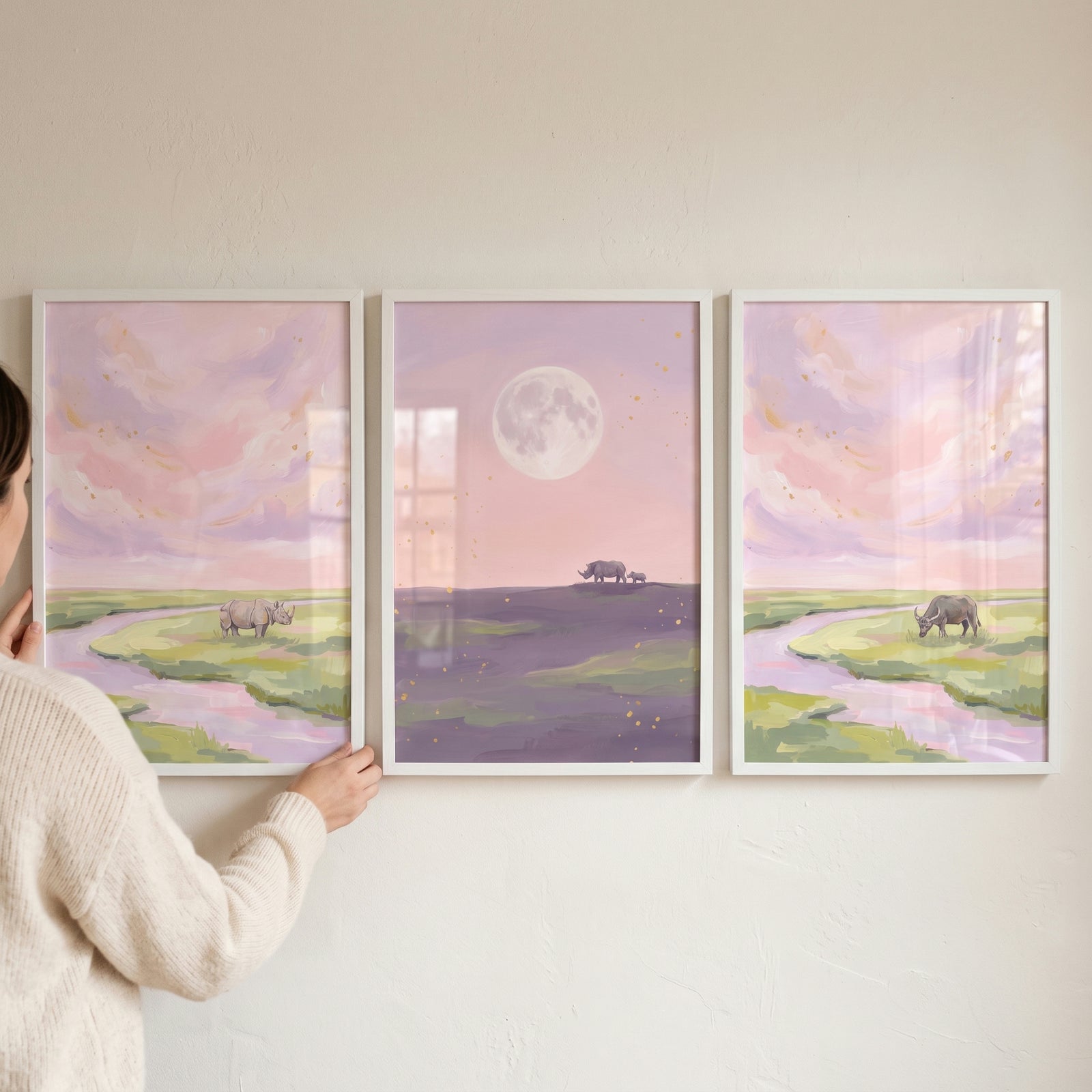 Woman holding a blush sky savanna animals wall art set of 3 featuring rhinos, a buffalo, and a moon.