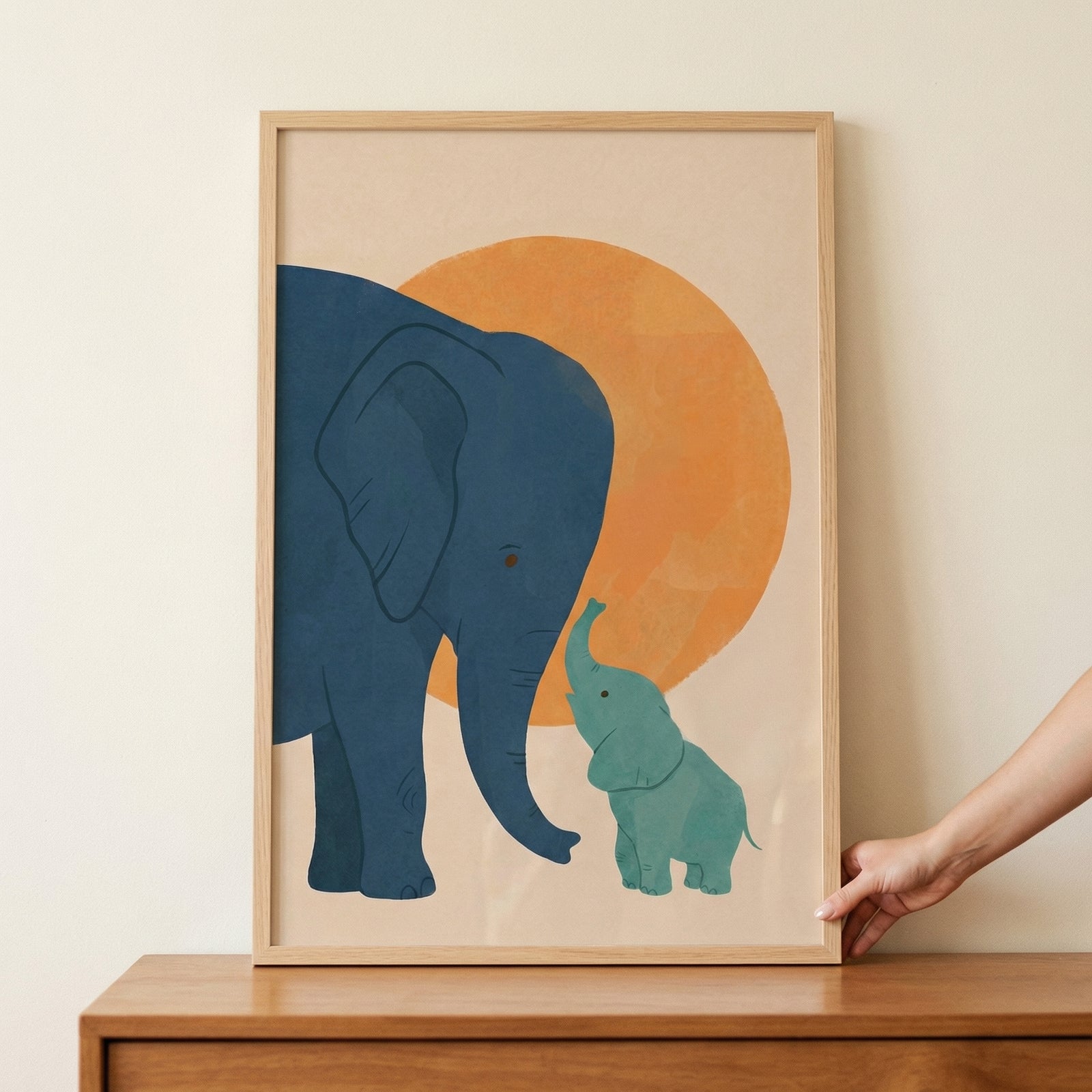 Framed illustration of a large blue elephant gently touching trunks with a smaller green elephant in front of a warm orange circle, resting on a wooden surface. The soft colors and simple shapes create a calm and nurturing feel suited for a child’s room.
