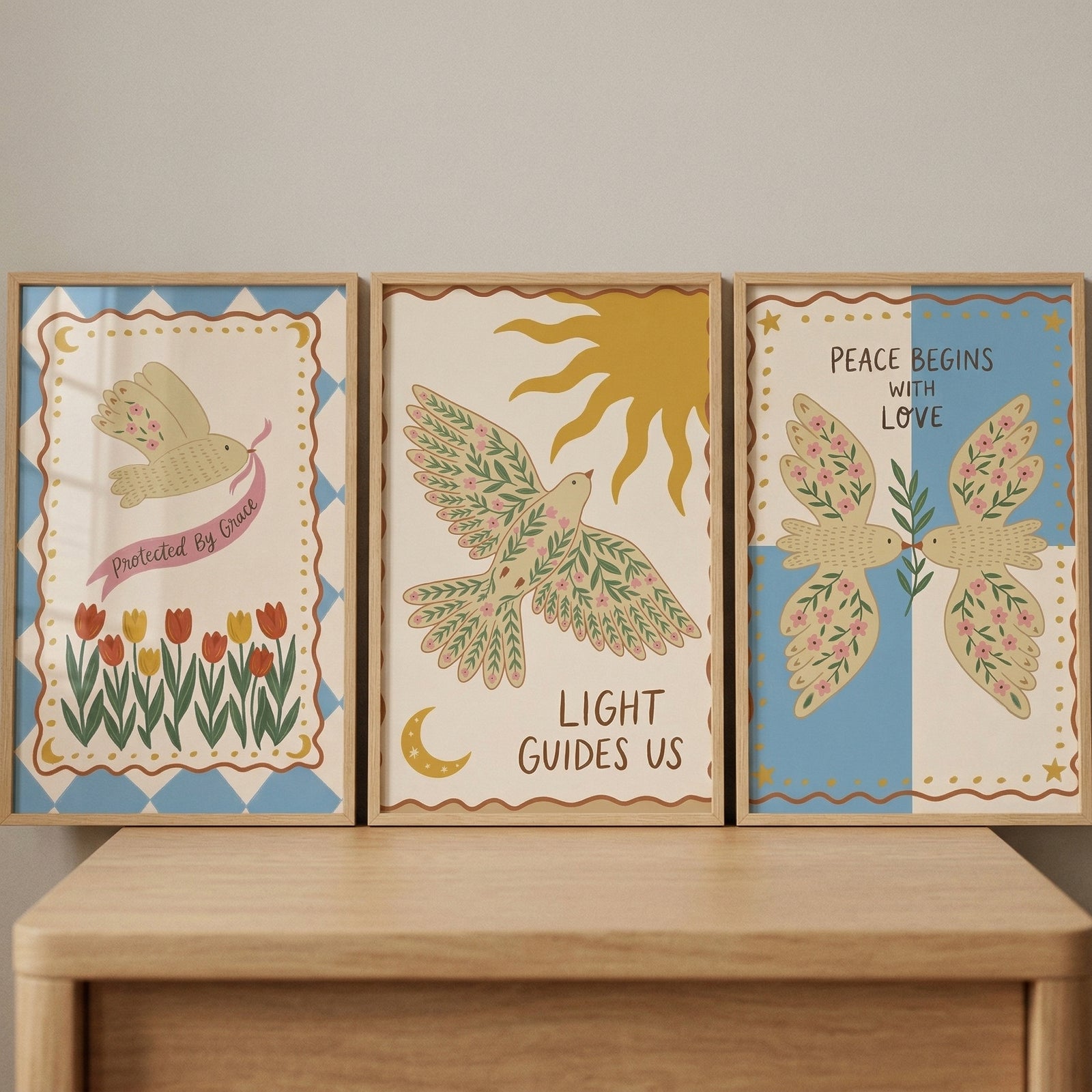 Three framed illustrations in a folk-art style are lined up on a light-colored wooden surface. Each piece features a cream-colored background with whimsical bird imagery and inspirational text.
