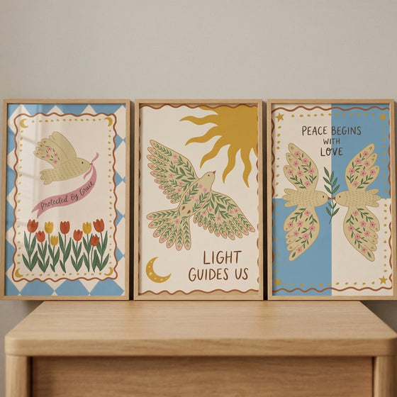 Three framed illustrations in a folk-art style are lined up on a light-colored wooden surface. Each piece features a cream-colored background with whimsical bird imagery and inspirational text.
