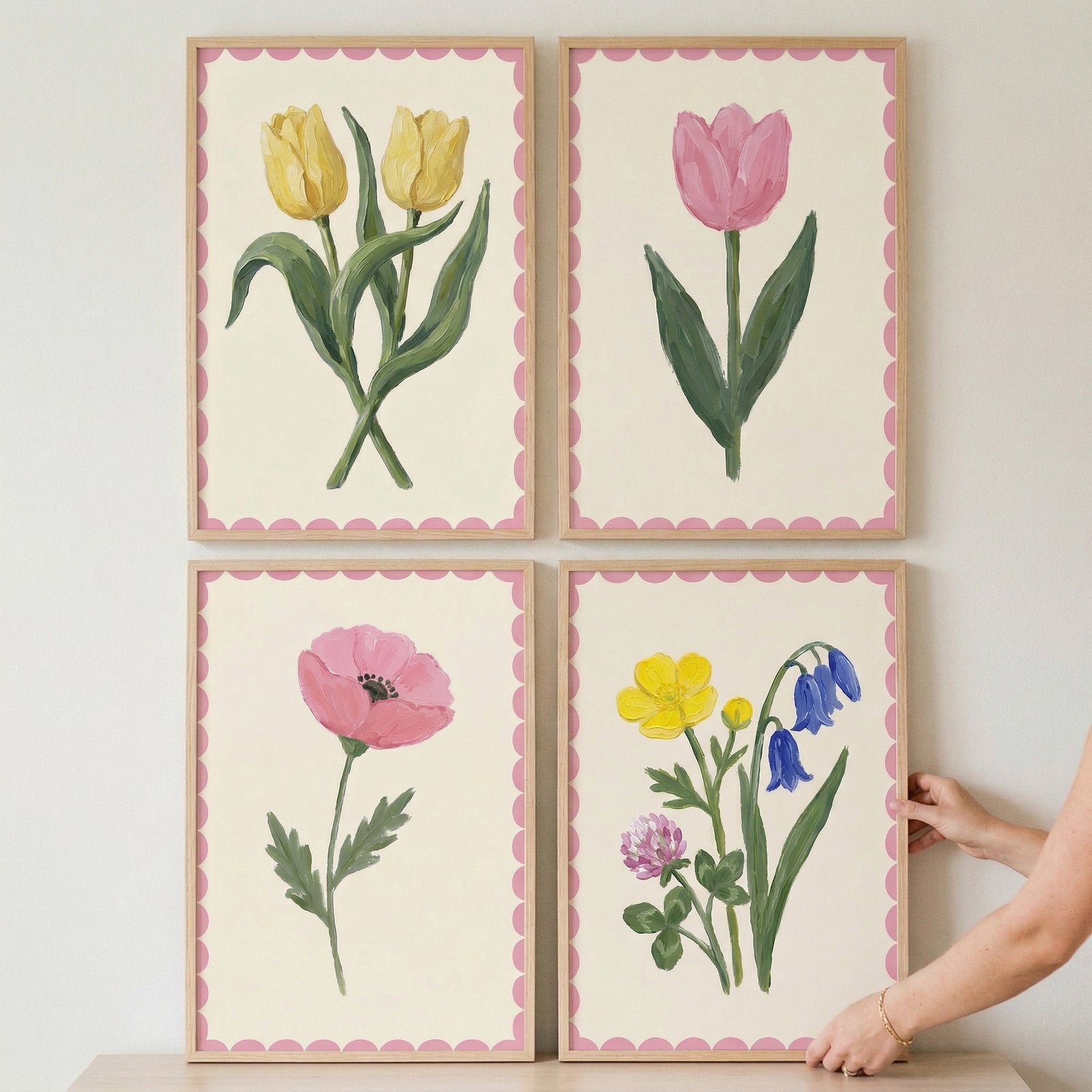 Four framed botanical prints arranged in a grid with pink scalloped borders, featuring yellow tulips, a pink tulip, a pink poppy, and a mix of small yellow, blue, and pink flowers. A hand adjusts the bottom right frame, suggesting the artwork being styled or arranged on a shelf or wall.