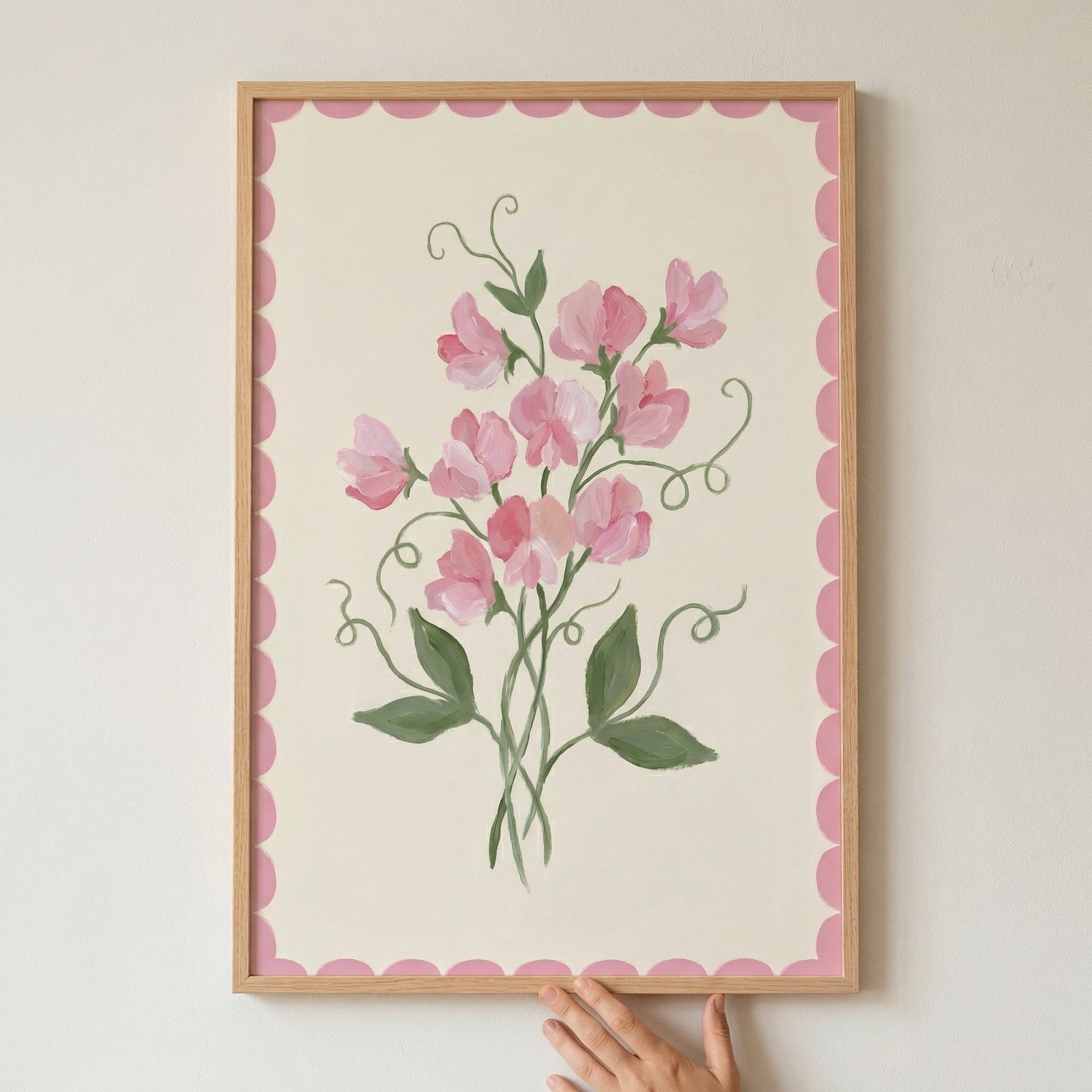 Framed painting of delicate pink flowers with soft green stems and curling tendrils set against a light background with a pink scalloped border. A hand gently holds the bottom of the frame, emphasizing the artwork’s soft and feminine floral detail.