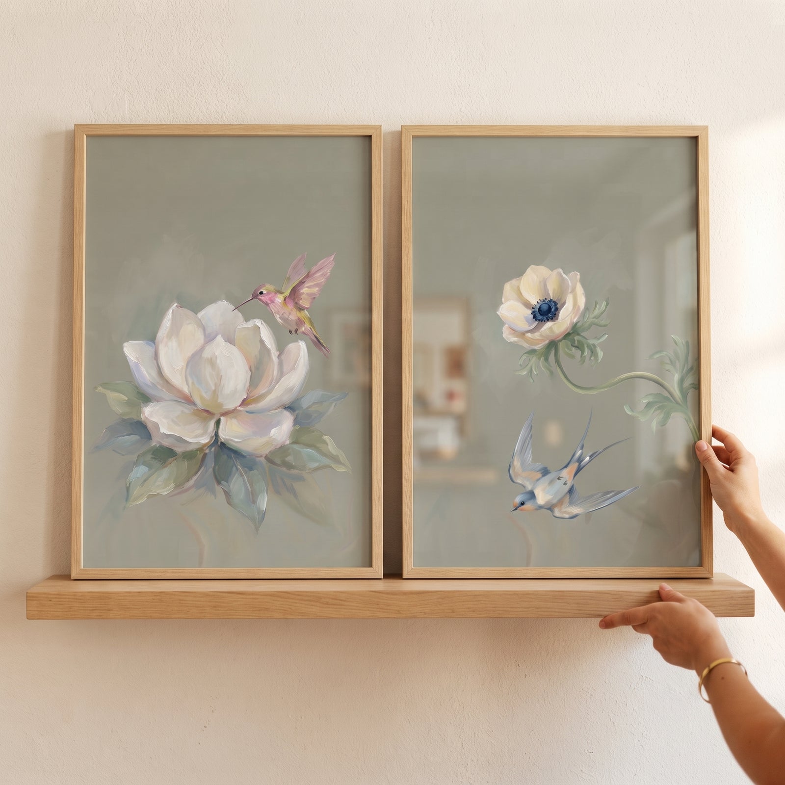 Hands hold a botanical white flower bird wall art set of 2 in light wood frames on a shelf.