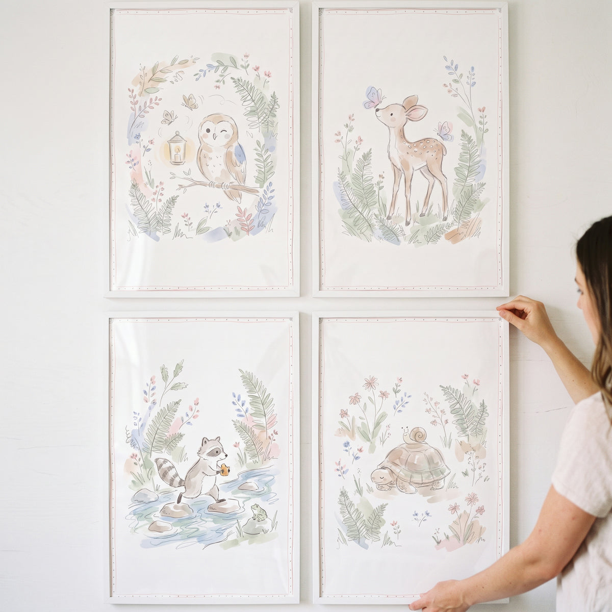 A person hangs a botanical wildflower woodland animals wall art set of 4 featuring an owl, deer, raccoon, and turtle.
