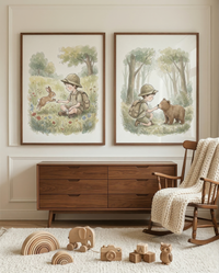Two framed watercolor-style illustrations hang on a neutral wall above a wooden dresser. The left art shows a boy in explorer clothes sitting in a meadow with a rabbit. The right art depicts him in a forest, kneeling to feed a small bear. To the right, a wooden rocking chair is draped with a cream knit blanket. On a white textured rug are various wooden toys, including a rainbow stacker, a toy elephant, a camera, and alphabet blocks.