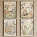 Four framed illustrations are arranged in a square grid on a neutral wall. Each piece features a gentle dinosaur family in a soft, whimsical storybook style. The artworks use muted earthy tones like tan, grey, and green with delicate floral and leaf accents. One shows a baby dinosaur holding a branch, another captures a family huddle, a third depicts a dinosaur hopping over a log, and the final scene shows two parents using a floral vine to swing their baby.