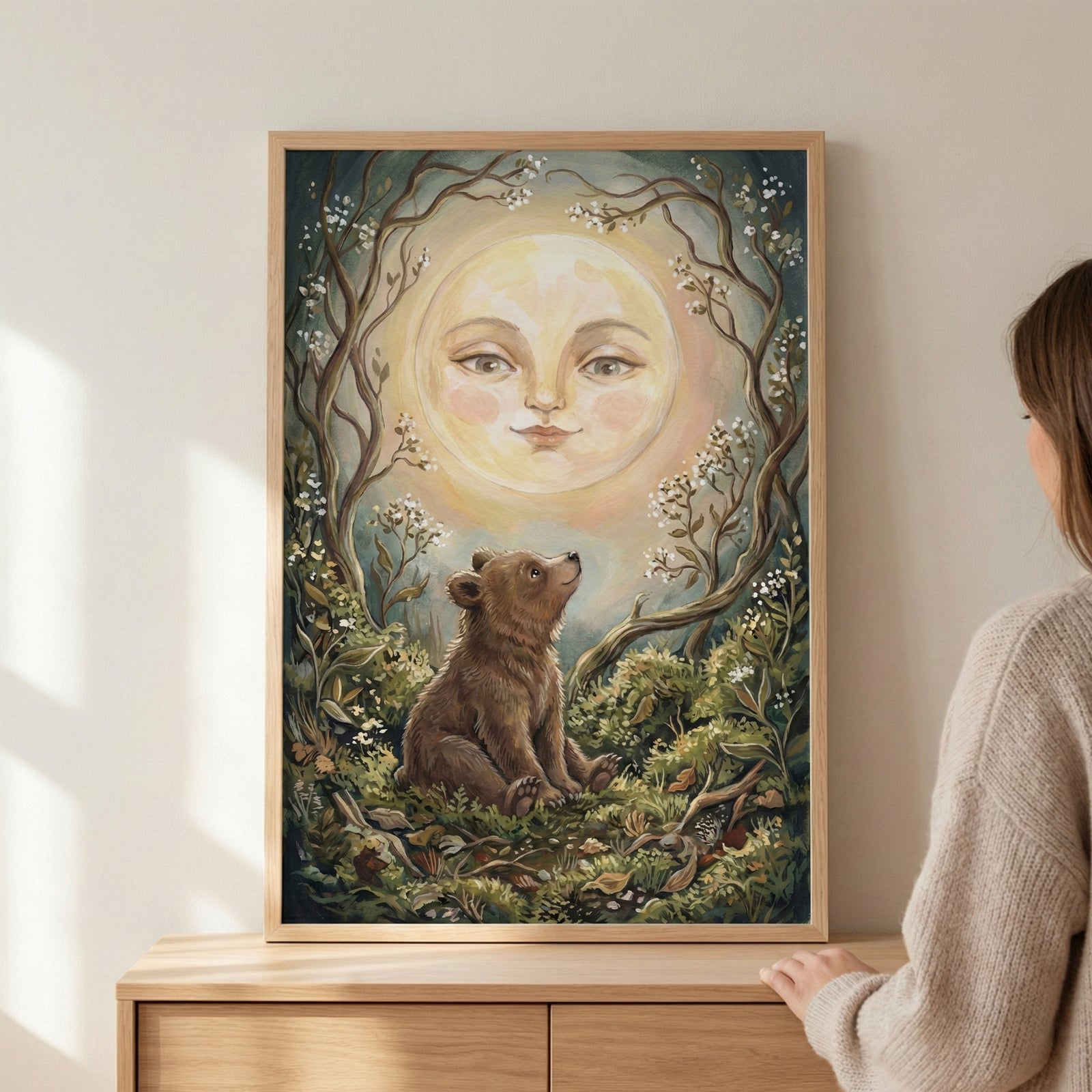 A framed piece of wall art featuring a young brown bear cub sitting on a forest floor, looking up at a large, glowing full moon with a serene, human-like face. The scene is framed by curved tree branches with small white blossoms and lush green foliage. To the right, the shoulder and back of a person in a light-colored sweater are visible as they look at the art, which is displayed on a wooden chest of drawers against a plain wall.