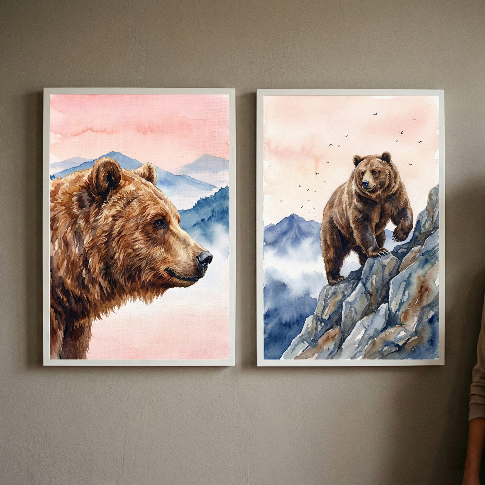 Two framed watercolor paintings of brown bears hanging on a neutral wall. The left painting shows a close up profile of a brown bear with soft pink sky and layered blue mountains behind it while the right painting shows a brown bear standing on a rocky cliff with misty mountains and small birds in the sky.