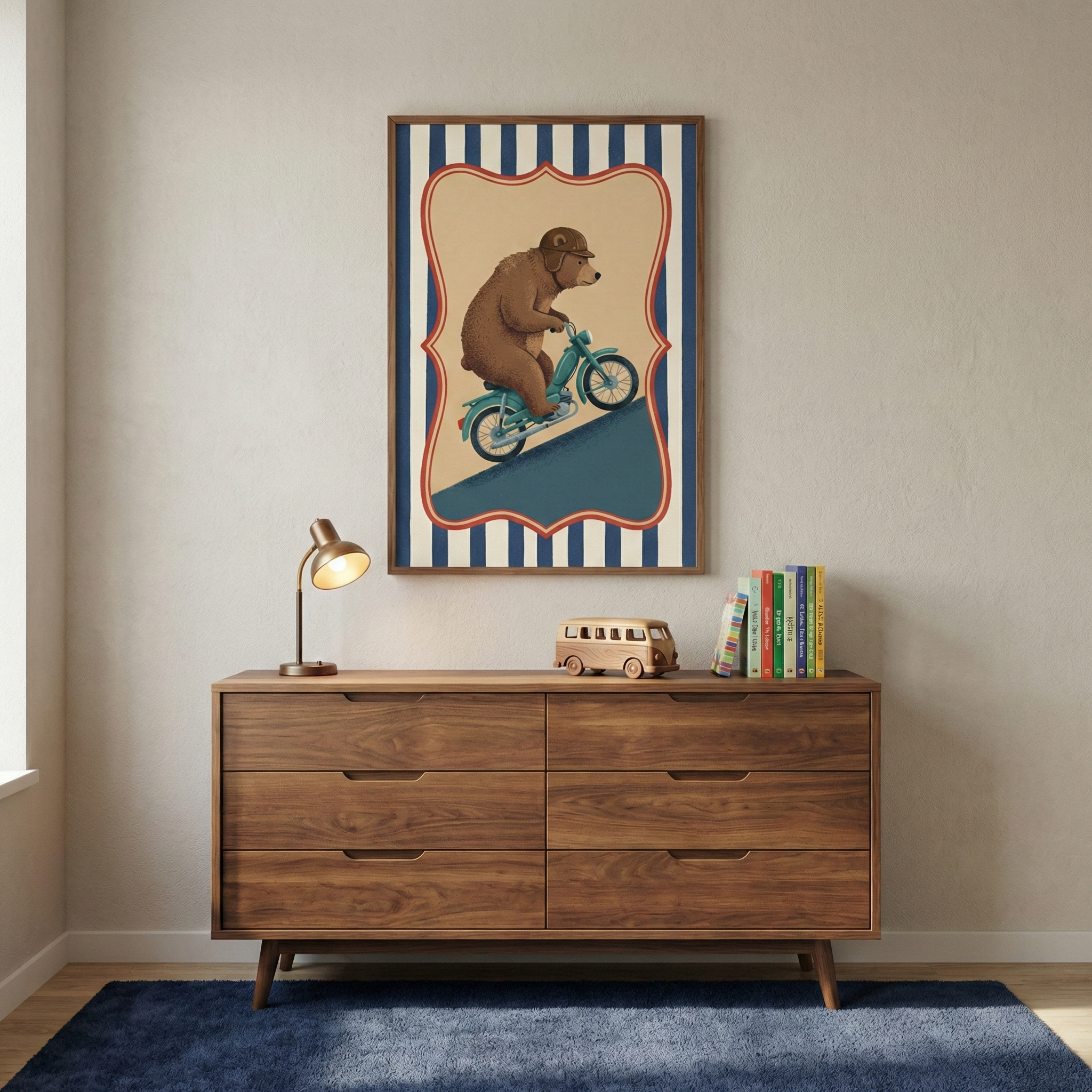 A framed piece of art hangs on a light-colored wall above a mid-century modern wooden dresser. The illustration features a brown bear wearing a vintage-style helmet and goggles while riding a light blue motorcycle up a hill, set against a background of blue and white vertical stripes with a decorative border. On top of the dresser, there is a small brass desk lamp with a warm glow, a simple wooden toy bus, and a small collection of colorful books. The dresser sits on a dark blue rug in a sunlit room.