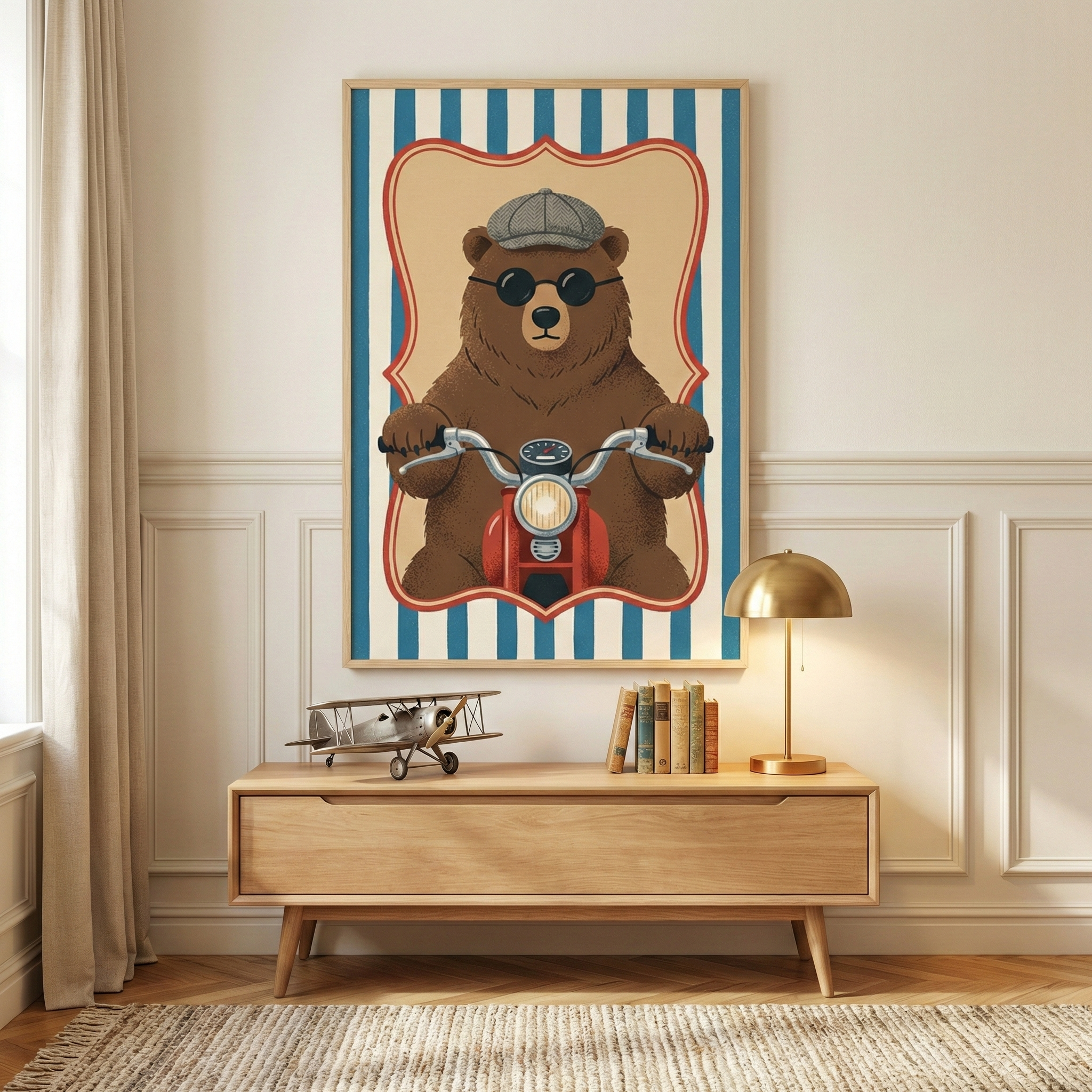A framed illustration hanging on a cream-colored wall with white wainscoting above a light wood console table. The central artwork features a large brown bear wearing round black sunglasses and a gray flat cap, riding a red vintage-style motorized scooter. The bear is centered within a cream-colored scalloped border against a background of vertical blue and white stripes. On the wooden table below the artwork sits a silver model airplane, a small stack of vintage books, and a lit brass table lamp with a dom