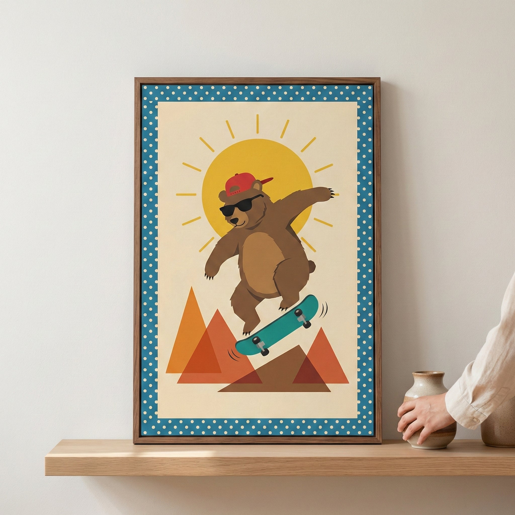 A framed graphic illustration of a cartoon brown bear performing a trick on a skateboard. The bear is depicted in a dynamic pose, wearing black sunglasses and a red baseball cap turned backward. Behind the bear is a large, radiant yellow sun and several sharp, stylized mountain peaks in shades of orange and brown. The image has a retro aesthetic with an off-white background and a decorative blue border with white polka dots. The artwork is set in a simple wooden frame and rests on a light wood shelf next to