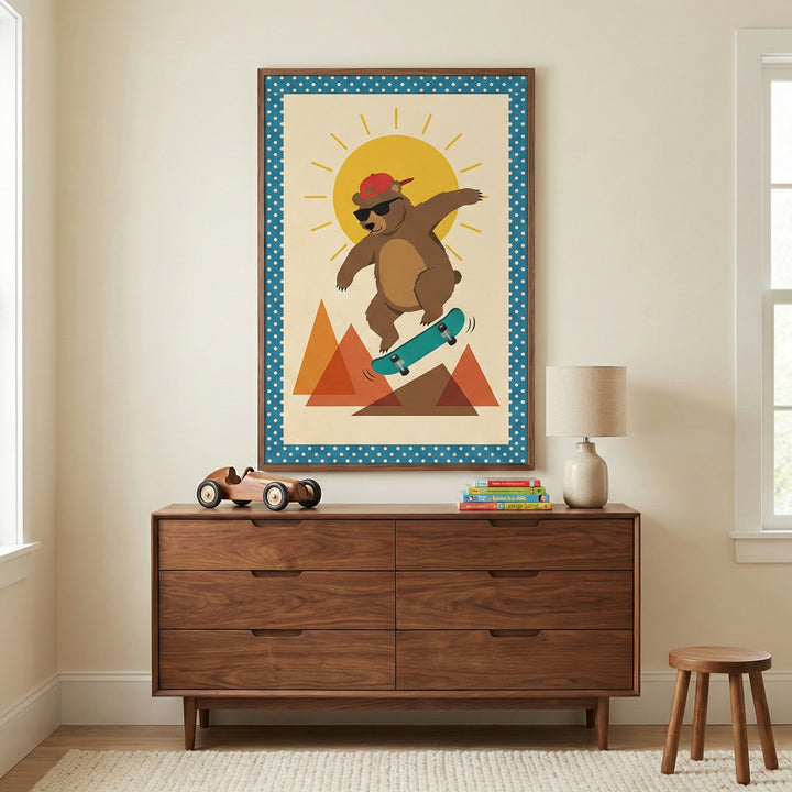 A framed illustration of a cartoon brown bear wearing a red baseball cap and sunglasses while skateboarding. The bear is depicted in front of a large sun and geometric orange mountains, surrounded by a blue polka-dot border. The art hangs on a cream wall above a mid-century style wooden dresser. On the dresser sits a wooden toy car, a small stack of books, and a lamp. A simple wooden stool is positioned on a textured rug in the corner of the room.