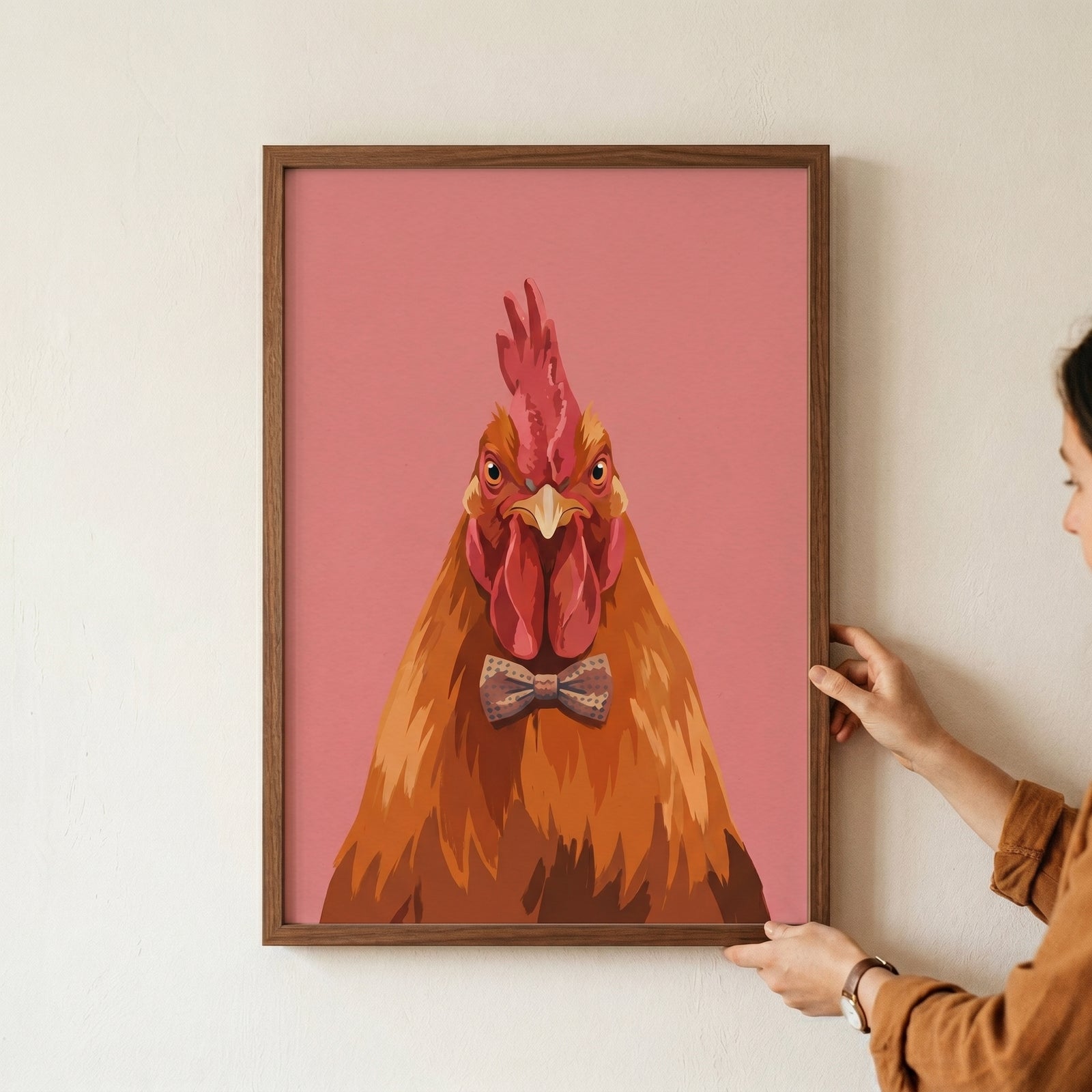 A person holds a framed brown bow tie rooster wall art print showing a dapper chicken portrait on a pink background.