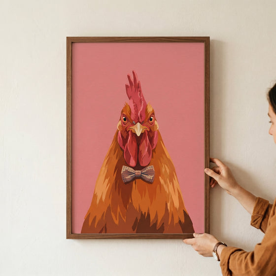 A person holds a framed brown bow tie rooster wall art print showing a dapper chicken portrait on a pink background.
