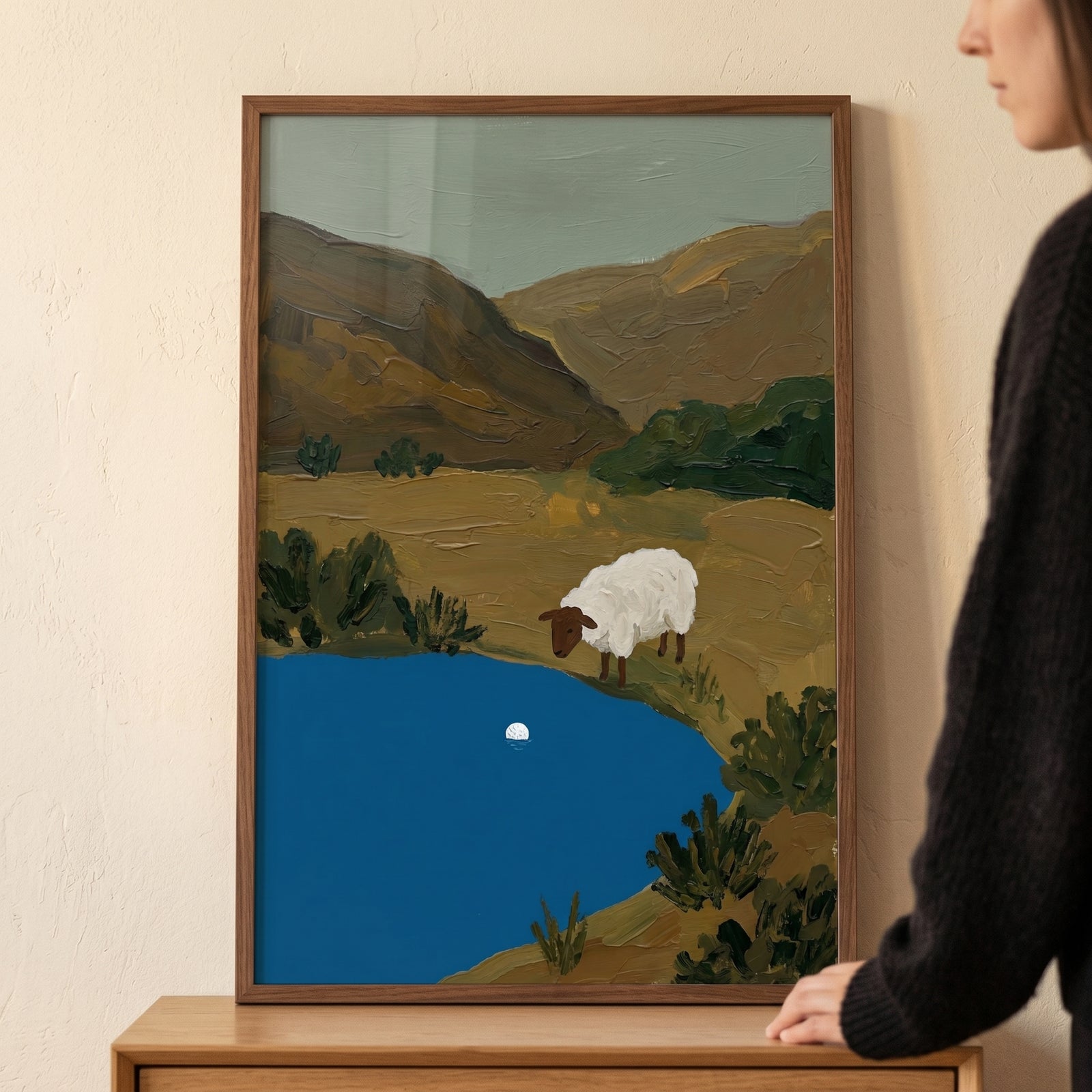 A white sheep stands by a blue pond in a hilly landscape in this framed brown face water sheep wall art.