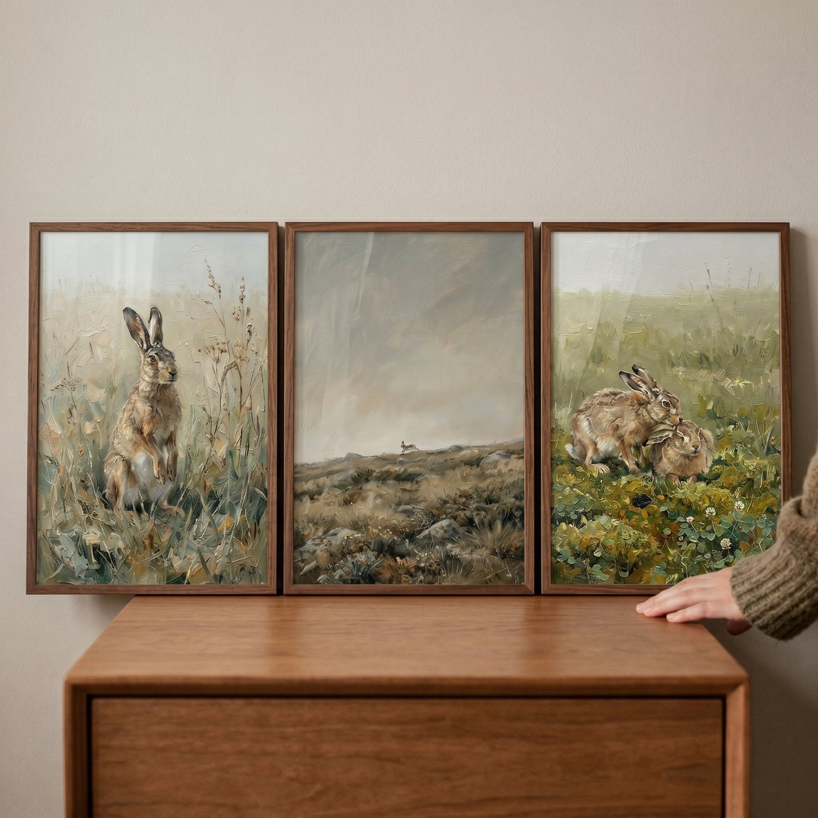 A hand touches a brown field hare wall art set of 3 in wood frames, displayed atop a smooth wooden dresser.