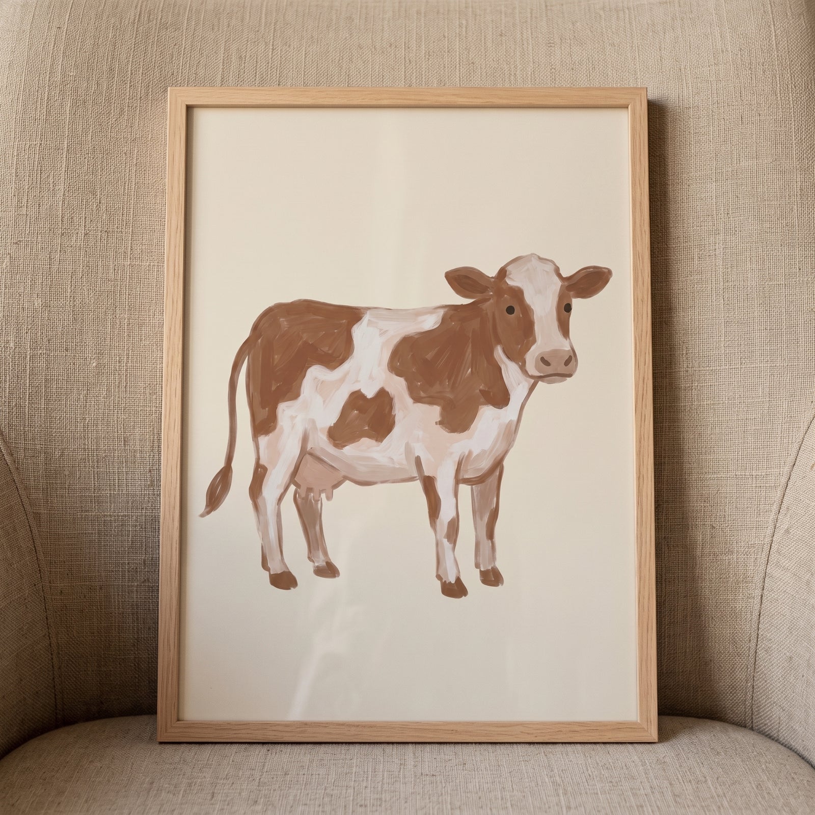 Brown white spotted cow wall art in a light wood frame rests on a textured beige armchair.