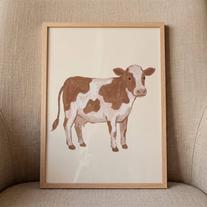 Brown white spotted cow wall art in a light wood frame rests on a textured beige armchair.
