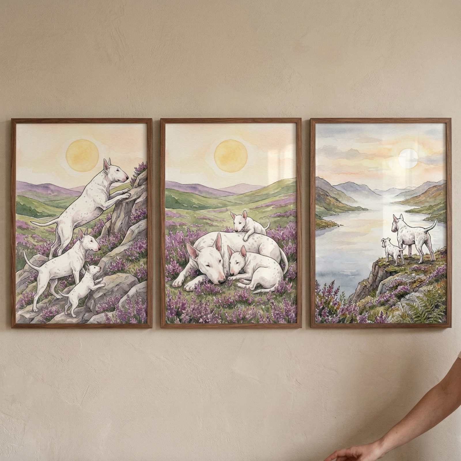 Three wooden-framed illustrations hang side-by-side on a light-colored wall, featuring white Bull Terriers in a soft watercolor style. The left image shows three dogs climbing a rocky slope with purple flowers. The middle image depicts a larger dog resting in a floral field with two small puppies. The right image shows two dogs on a cliff overlooking a misty lake and mountains. Each scene is set under a soft, glowing sun against a background of rolling green hills in muted tones.