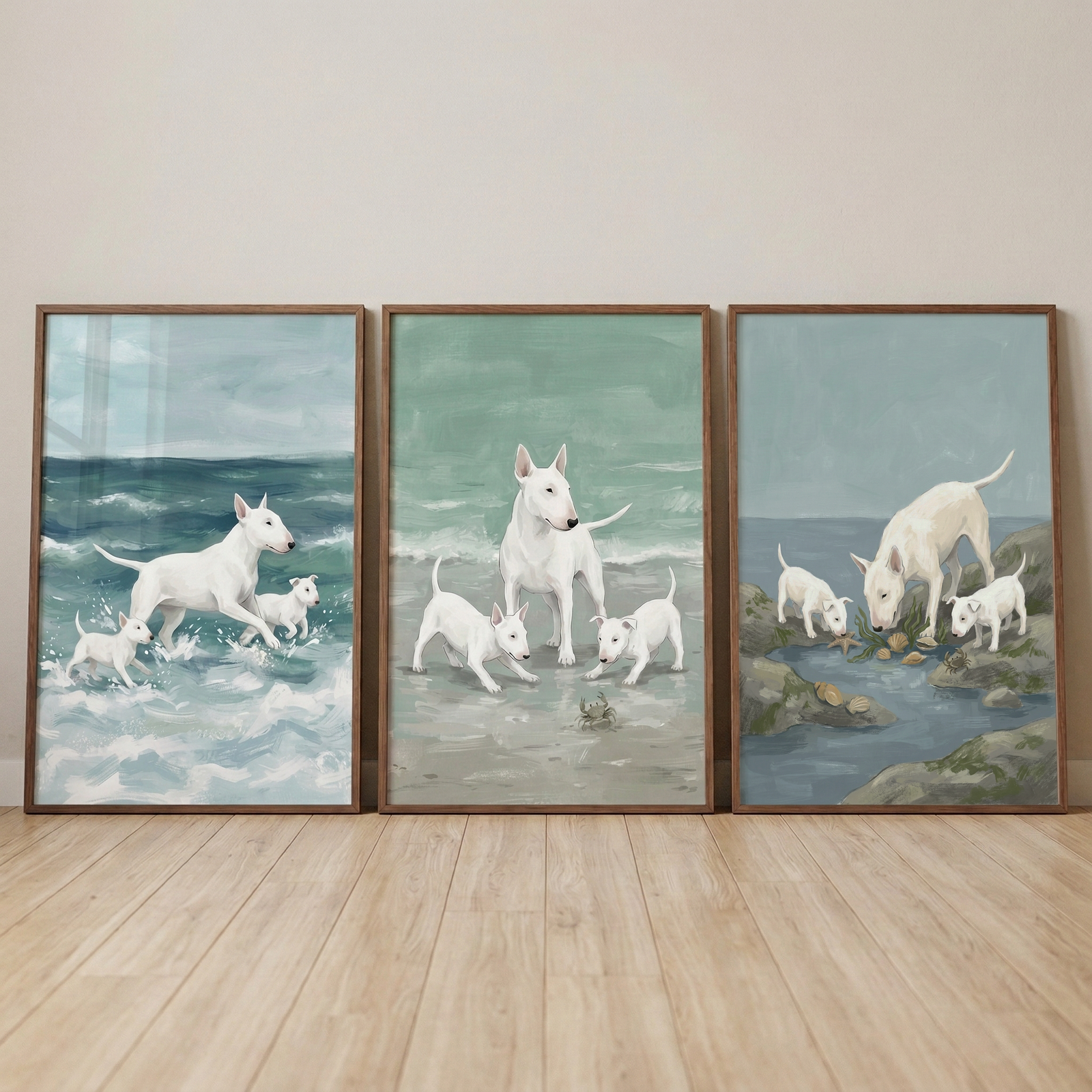 Three framed pieces of art leaning against a neutral wall on a light wood floor. Each piece features white bull terriers in coastal settings. On the left, an adult dog and two puppies run through blue ocean waves. The center scene shows the dogs on a sandy beach near a small crab. The right scene depicts them exploring rocks and tide pools. The style uses soft brushwork and a muted color palette of seafoam green and ocean blue.