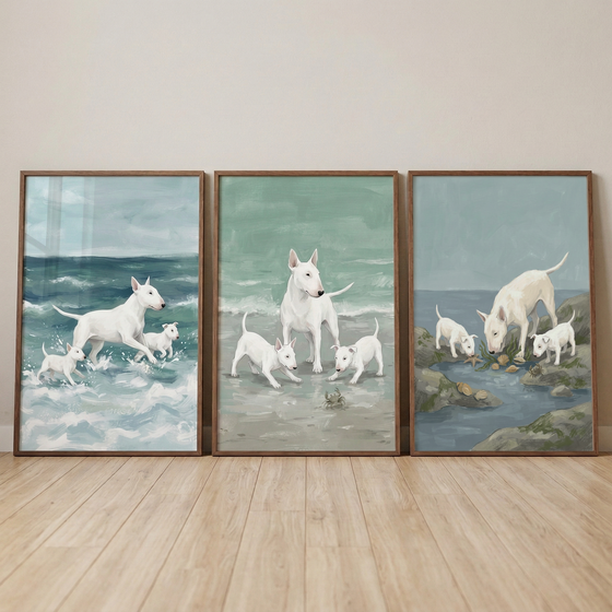 Three framed pieces of art leaning against a neutral wall on a light wood floor. Each piece features white bull terriers in coastal settings. On the left, an adult dog and two puppies run through blue ocean waves. The center scene shows the dogs on a sandy beach near a small crab. The right scene depicts them exploring rocks and tide pools. The style uses soft brushwork and a muted color palette of seafoam green and ocean blue.