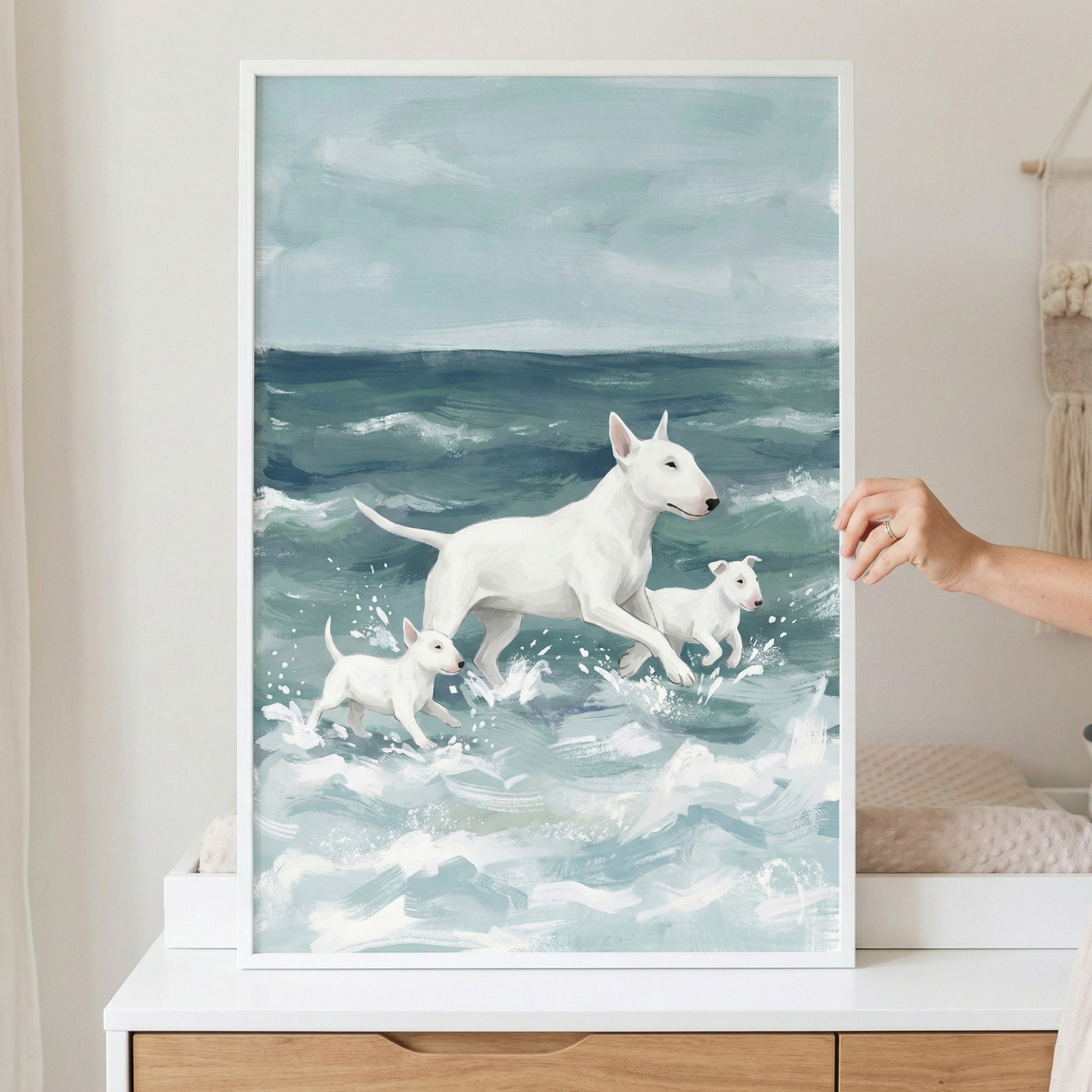 A vertically oriented framed illustration shows an adult white bull terrier and two small puppies wading through churning ocean waves. The large dog is in the center, while the smaller pups flank it, all facing to the right. The water is rendered with broad strokes of white foam over a teal sea. Above, a pale, cloudy sky completes the scene. The artwork, held by a hand, features a textured, expressive style and is enclosed in a white frame resting on a wooden cabinet.