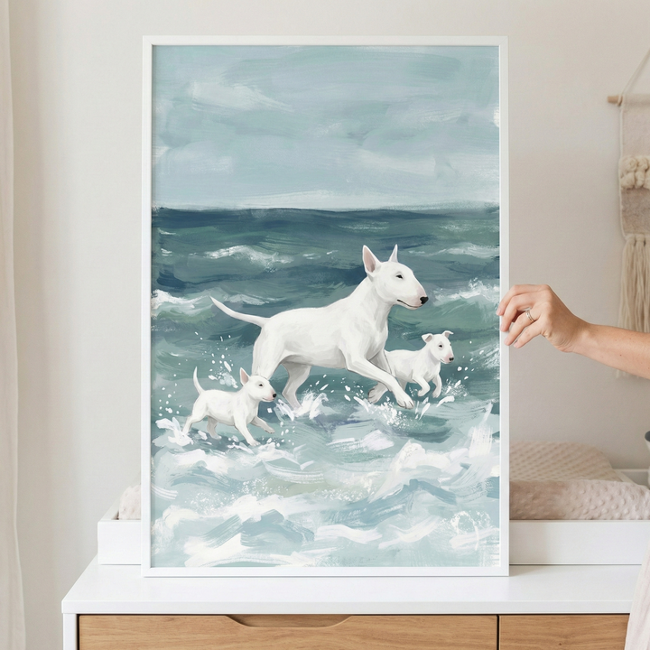 A vertically oriented framed illustration shows an adult white bull terrier and two small puppies wading through churning ocean waves. The large dog is in the center, while the smaller pups flank it, all facing to the right. The water is rendered with broad strokes of white foam over a teal sea. Above, a pale, cloudy sky completes the scene. The artwork, held by a hand, features a textured, expressive style and is enclosed in a white frame resting on a wooden cabinet.