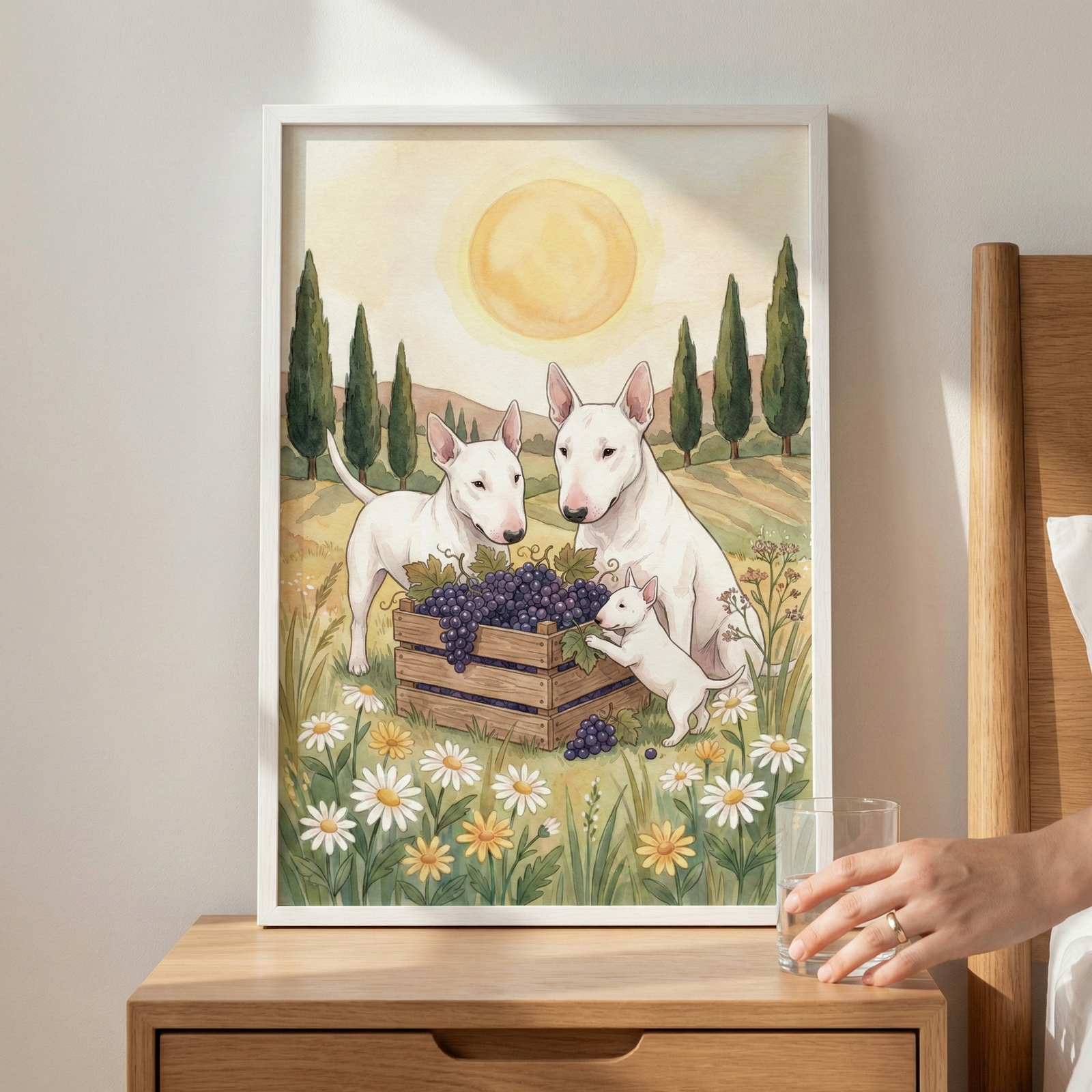 A vertical, white-framed illustration sits on a light-toned wooden nightstand next to a bed. The artwork depicts three white bull terrier dogs—two adults and a smaller puppy—in a sunlit meadow. They are gathered around a wooden crate overflowing with dark purple grapes. The foreground is filled with green grass and various white and yellow wildflowers. Behind the dogs, the rolling landscape features tall, slender trees under a large, pale yellow sun. To the right, a hand reaches for a glass of water.
