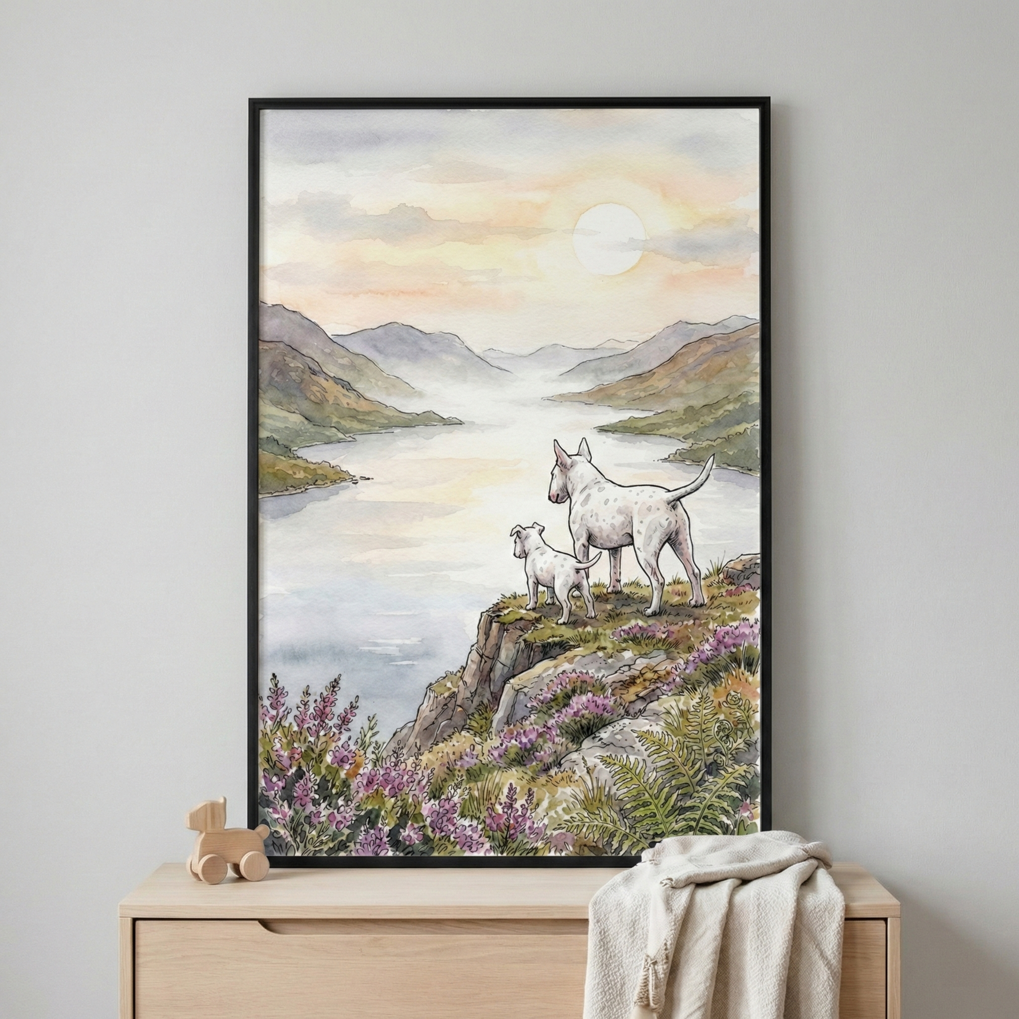 A vertical framed illustration in a watercolor style depicts two white dogs standing on a rocky cliffside. The adult dog and a smaller puppy gaze out over a calm body of water that winds between misty, layered mountains under a soft, hazy sky with a pale sun. The cliff is decorated with clusters of purple wildflowers and green ferns. The artwork, set in a slim black frame, hangs on a light gray wall above a wooden dresser. A small wooden toy and a folded white cloth sit on the dresser's surface.