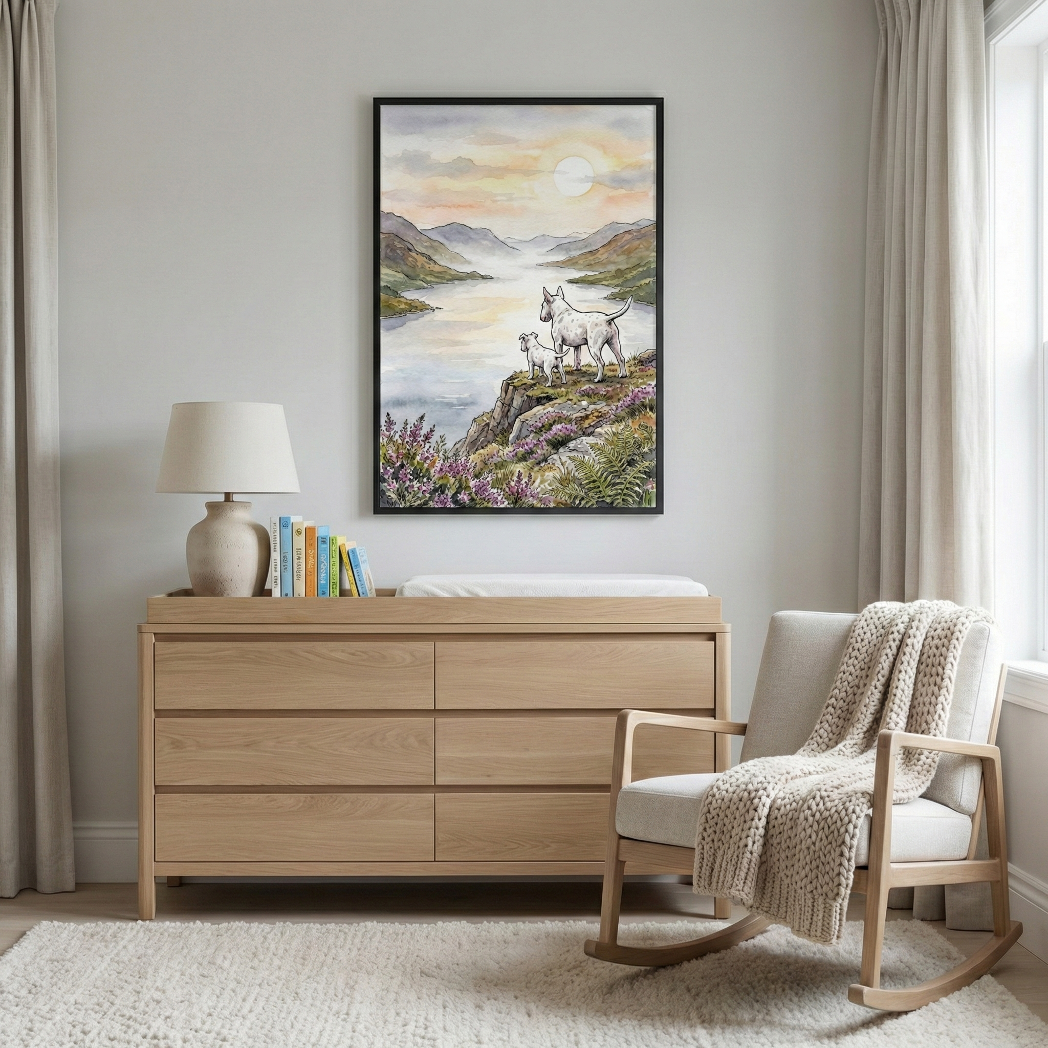 A serene nursery or bedroom setting with a light wood dresser as the centerpiece. On top of the dresser is a simple lamp with a textured base, a small collection of colorful books, and a white cushioned pad. A large, black-framed artwork hangs on the wall above, showing two white dogs on a rocky outcrop looking over a vast lake and distant mountains at dawn. The foreground of the artwork features purple flowers and green foliage. 